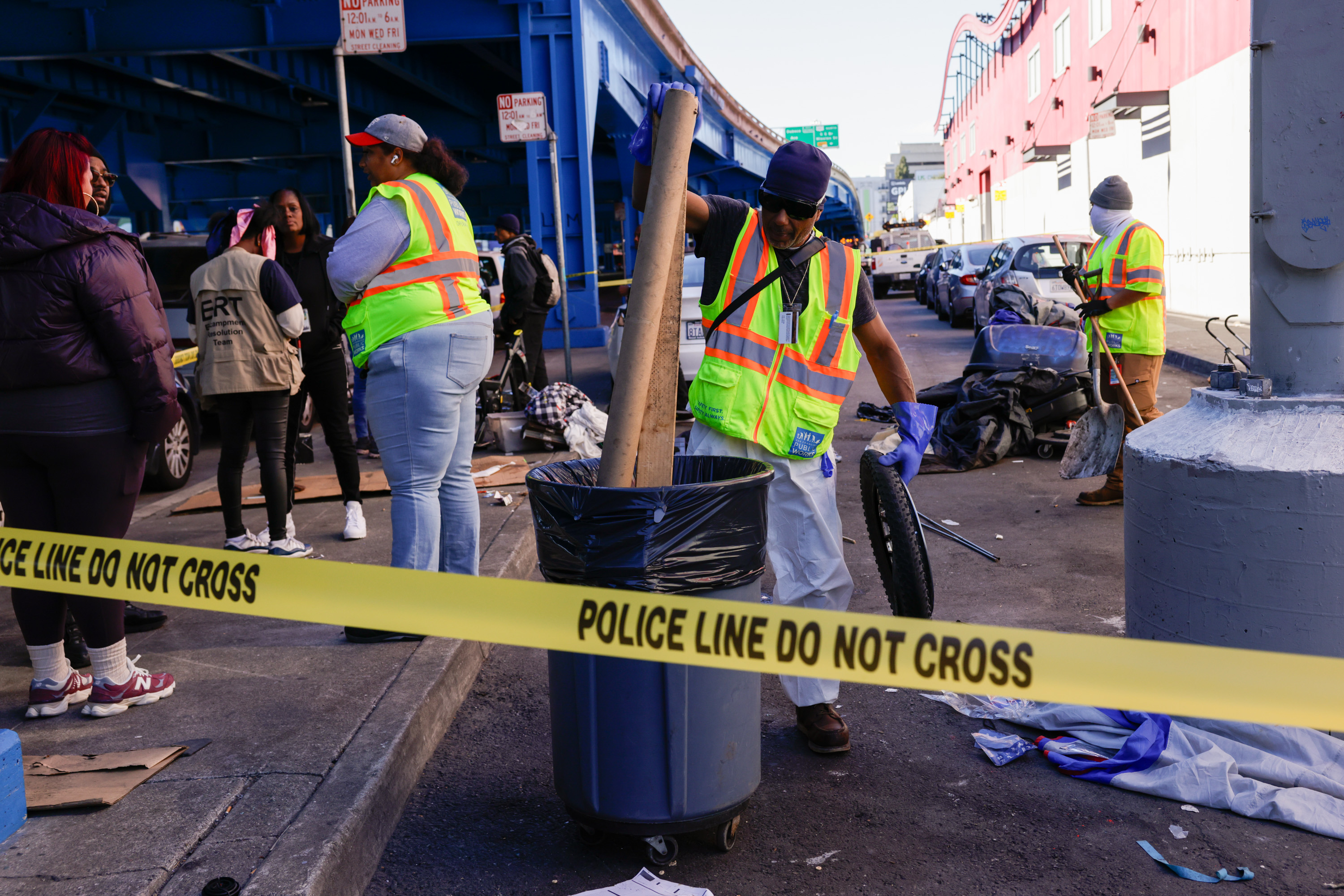 People in safety vests clean a street behind yellow police tape. They're near a blue structure, and various items are scattered on the ground.