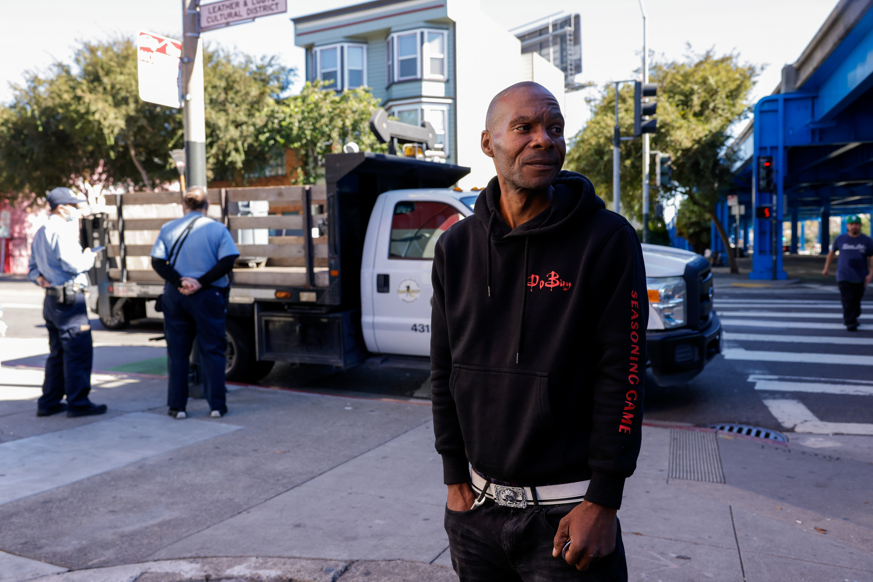 A man in a black hoodie stands on a street corner, hands in pockets. Behind him, two men stand near a truck, and buildings and trees are visible.