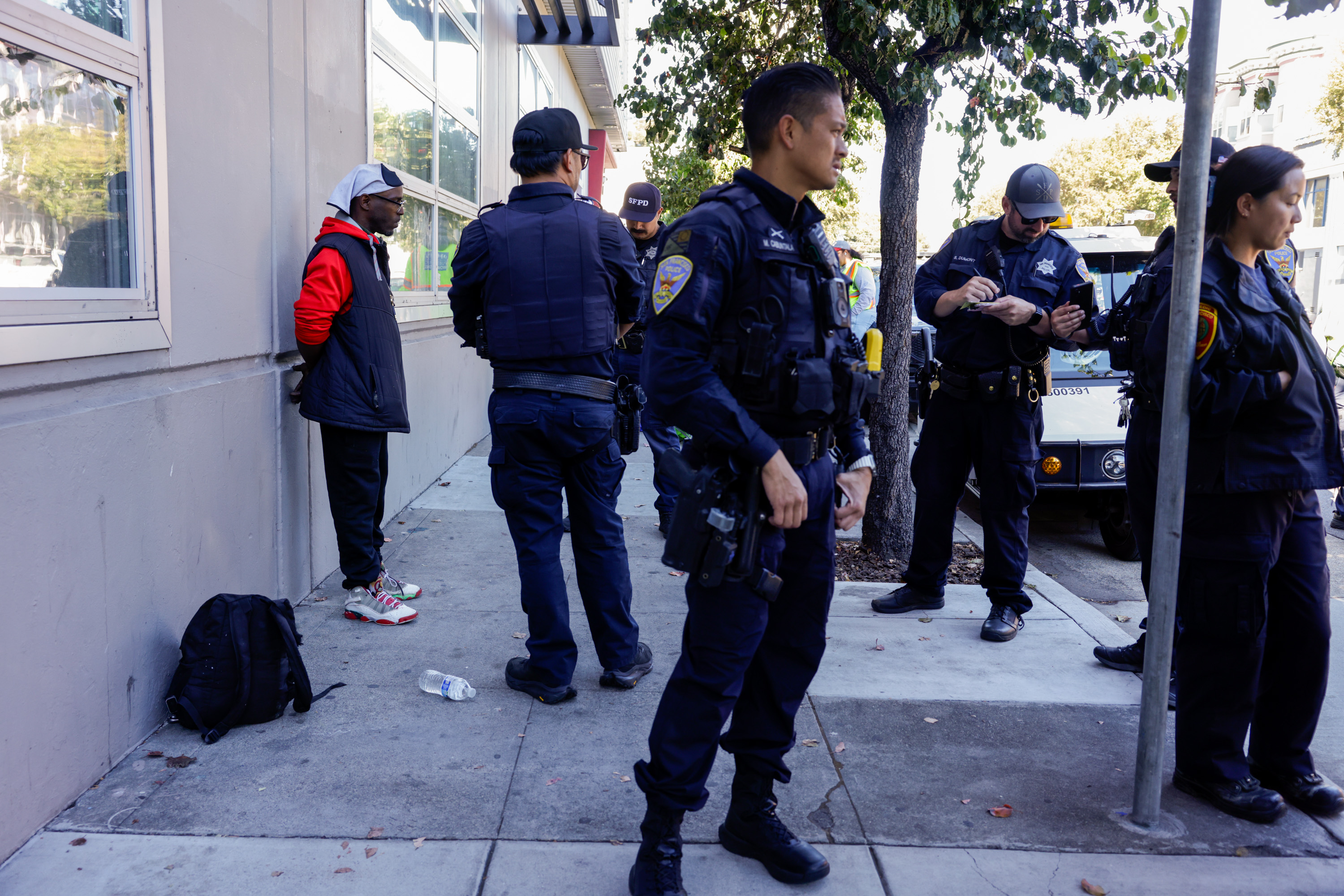 A man in a red and black jacket stands against a wall with his hands behind his back. Several police officers stand nearby, and a backpack and water bottle are on the ground.