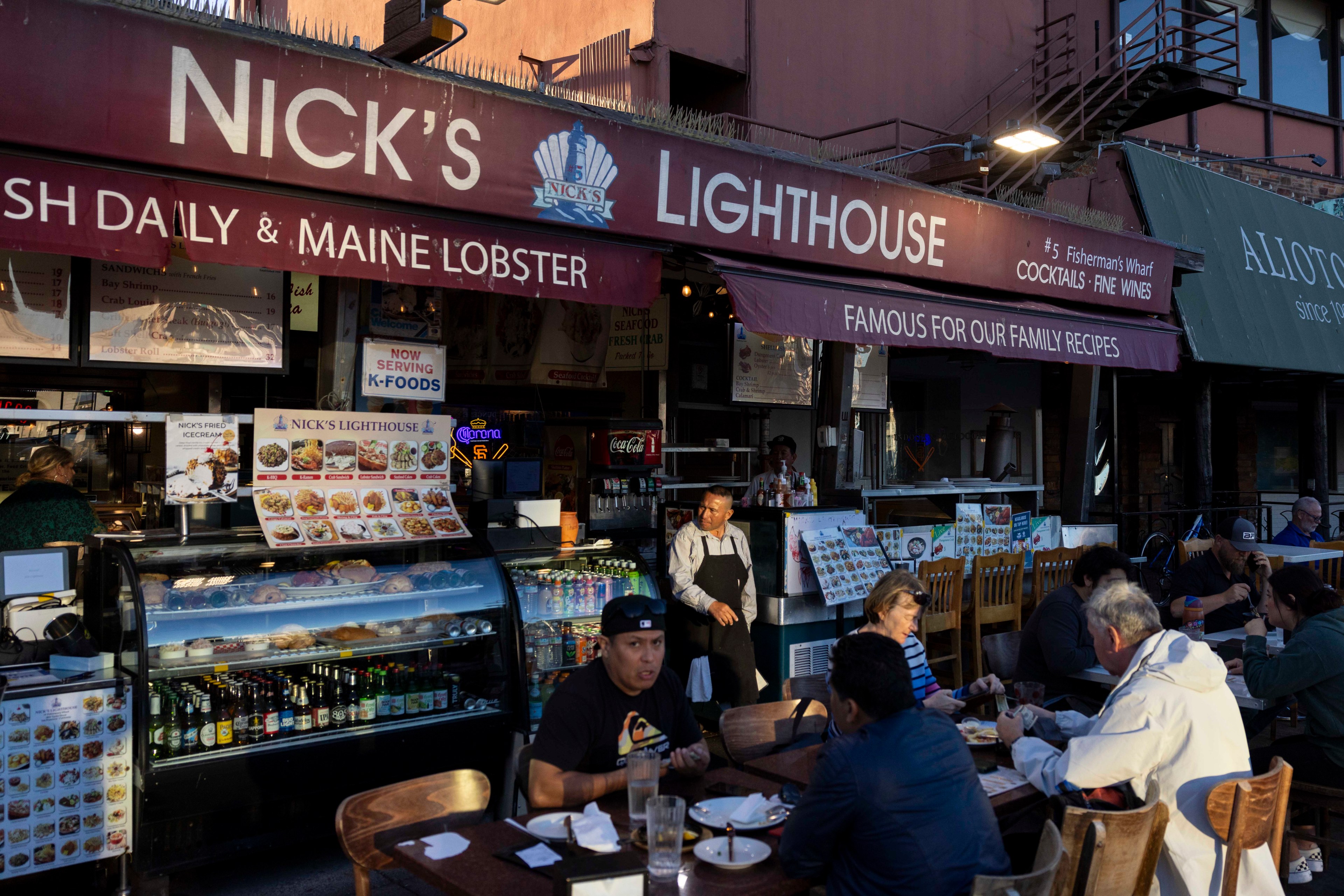 Outdoor dining at Nick's Lighthouse features a counter displaying seafood under a maroon awning. People sit at wooden tables enjoying their meals.