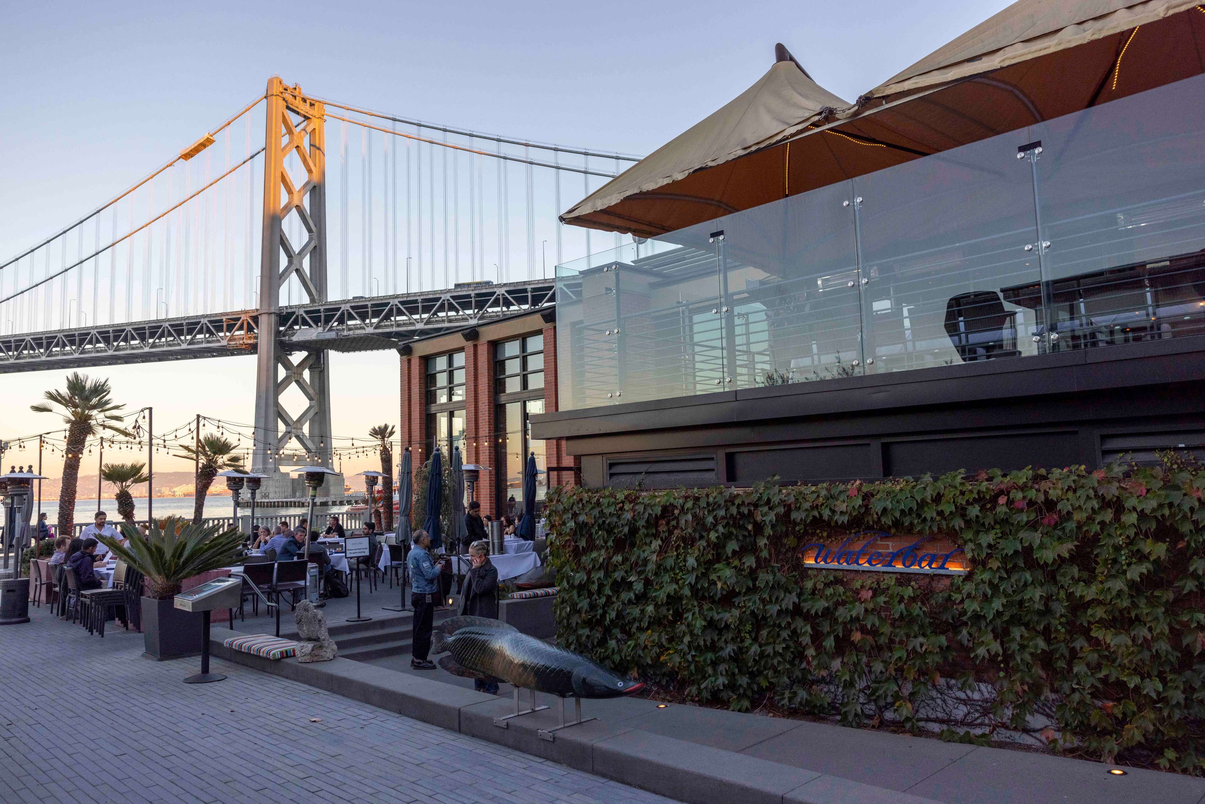 A bustling waterfront scene with a large bridge in the background, outdoor dining, palm trees, a structure with a canopy, and a decorative fish sculpture.