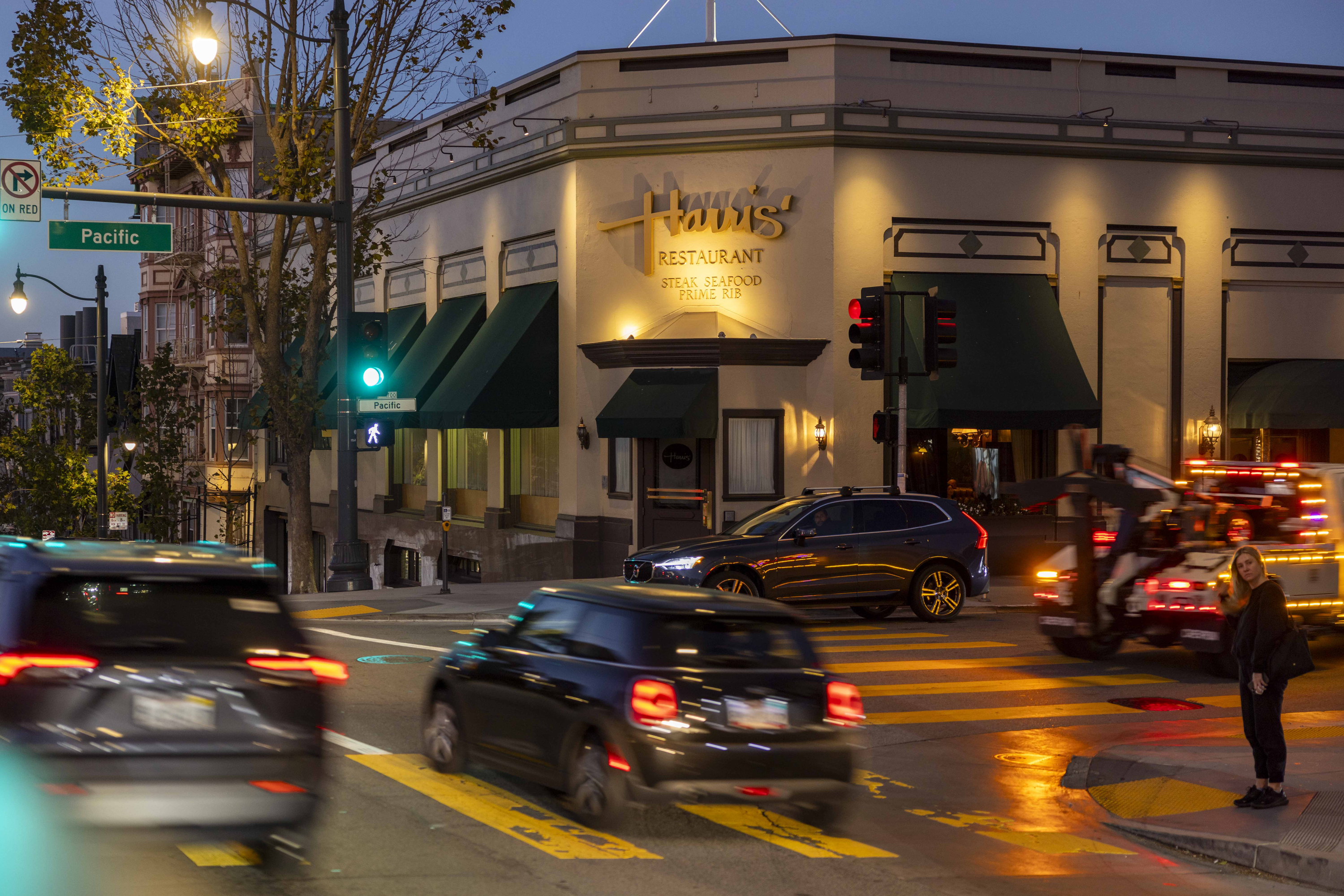 A busy intersection at dusk with blurred cars and a woman standing on the corner. A restaurant named Harris' is lit up, with green awnings visible.