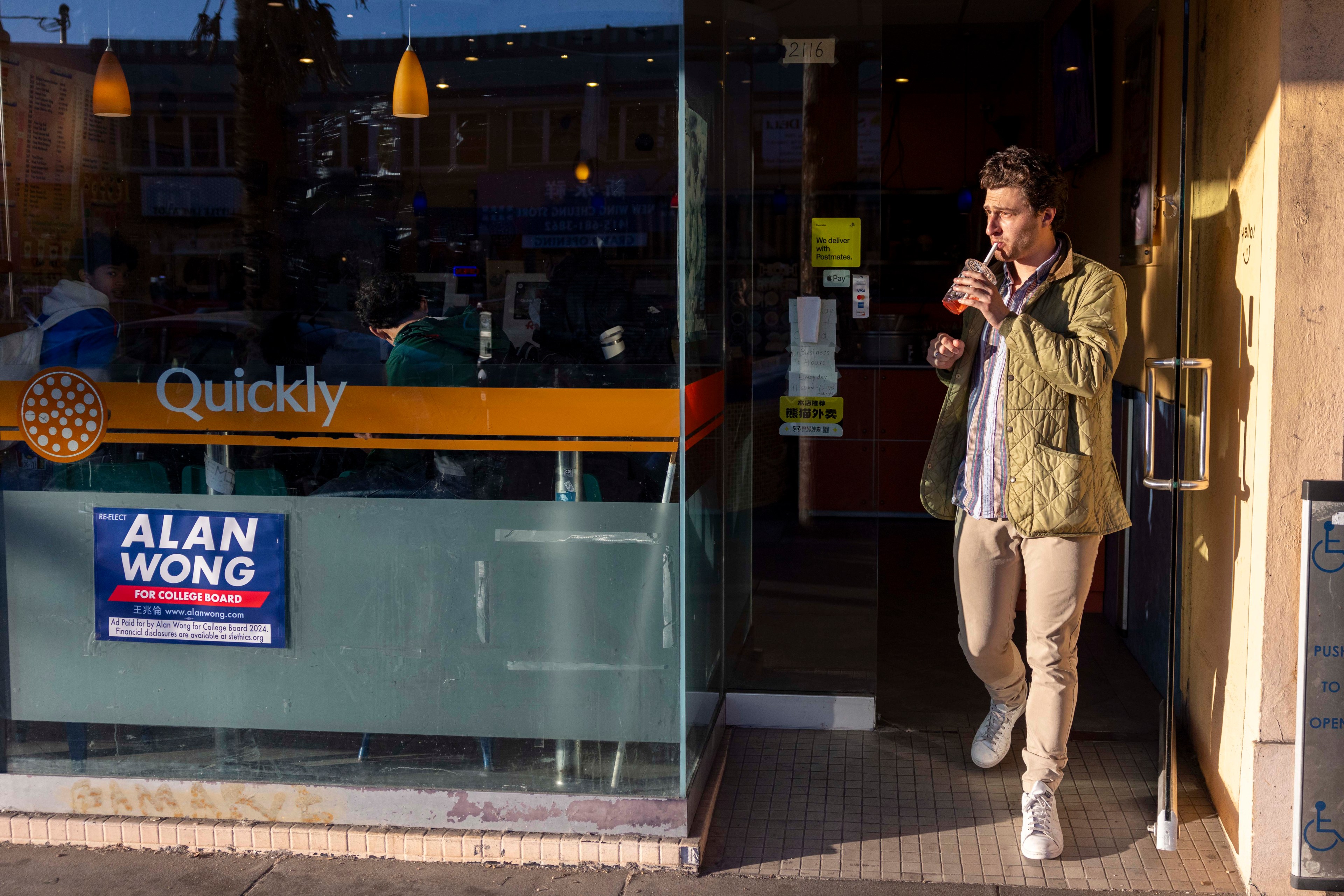 A man in a green jacket is drinking from a cup while exiting a bubble tea shop named Quickly. A campaign sign for Alan Wong is on the window.