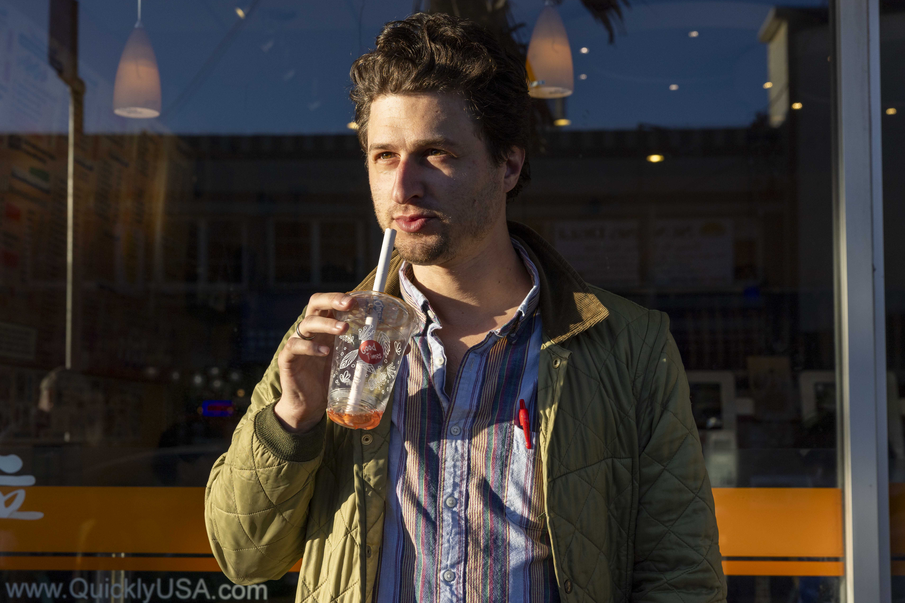 A person stands outside, sipping a drink through a straw from a cup. They wear a quilted jacket over a striped shirt, with pens visible in the shirt pocket.