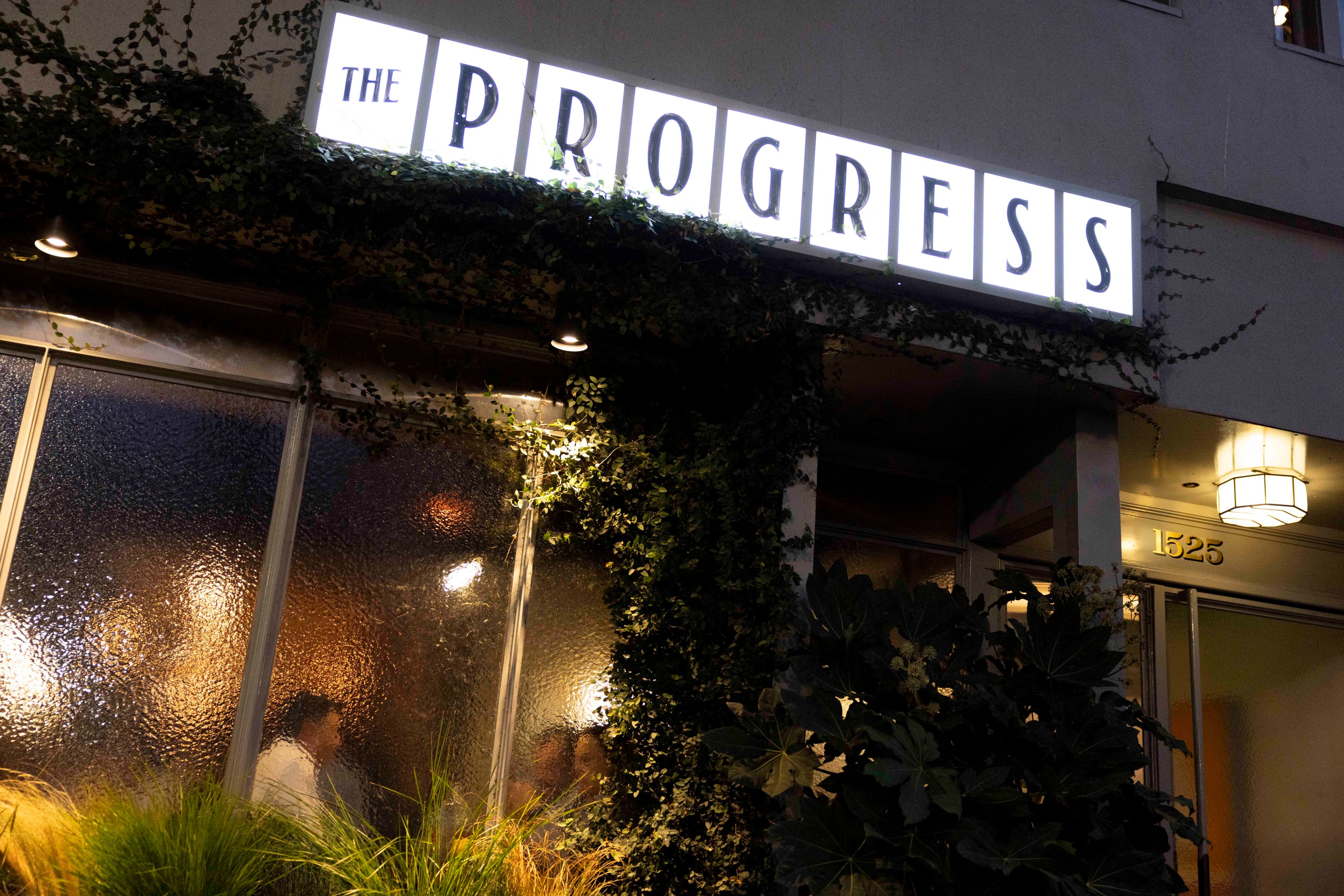 A restaurant entrance is shown with the sign &quot;THE PROGRESS&quot; illuminated above. The exterior is adorned with lush green plants, and the number 1525 is visible.