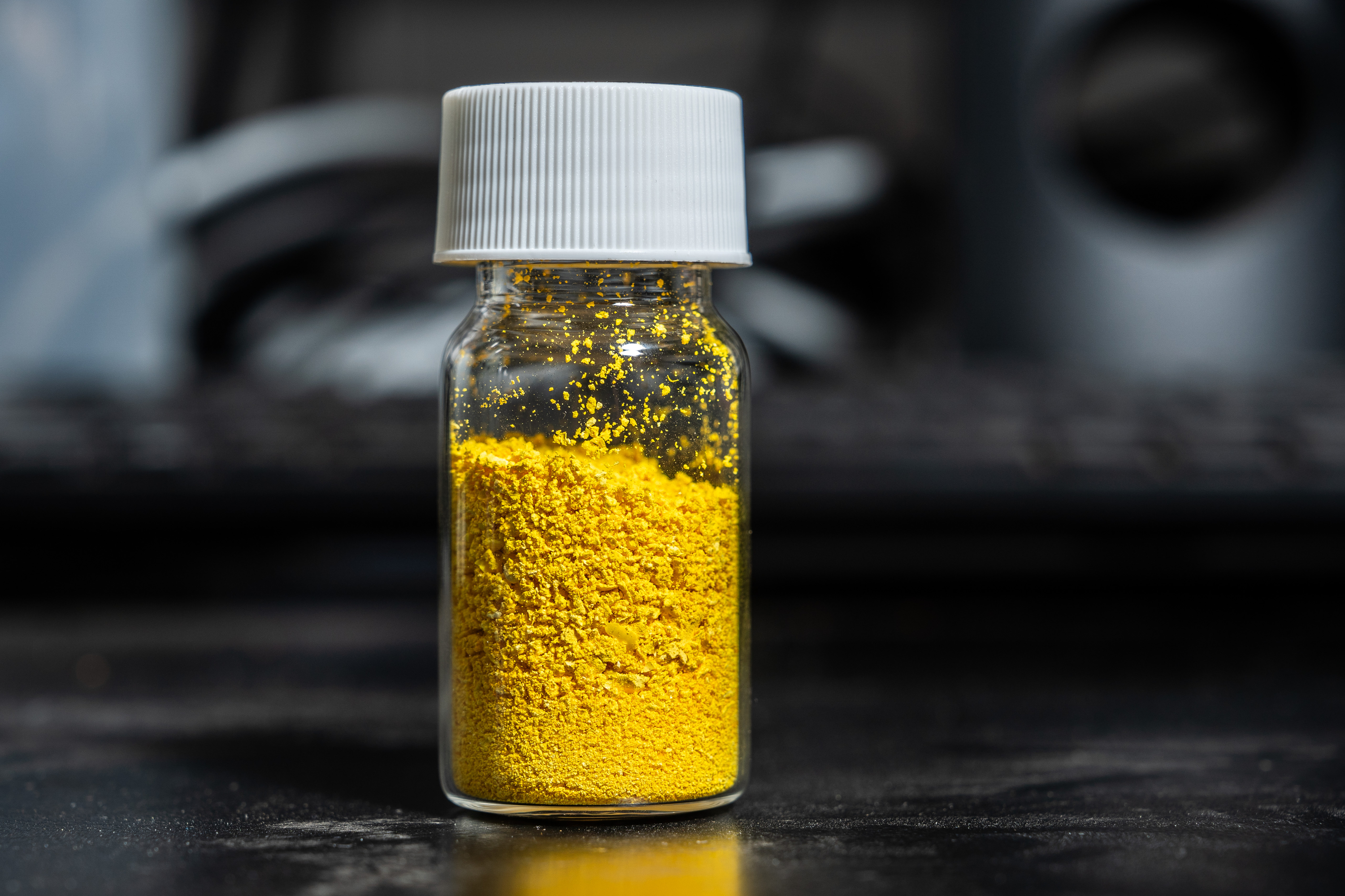 A small, clear glass jar with a white screw cap contains bright yellow powder, placed on a dark surface with a blurred background.