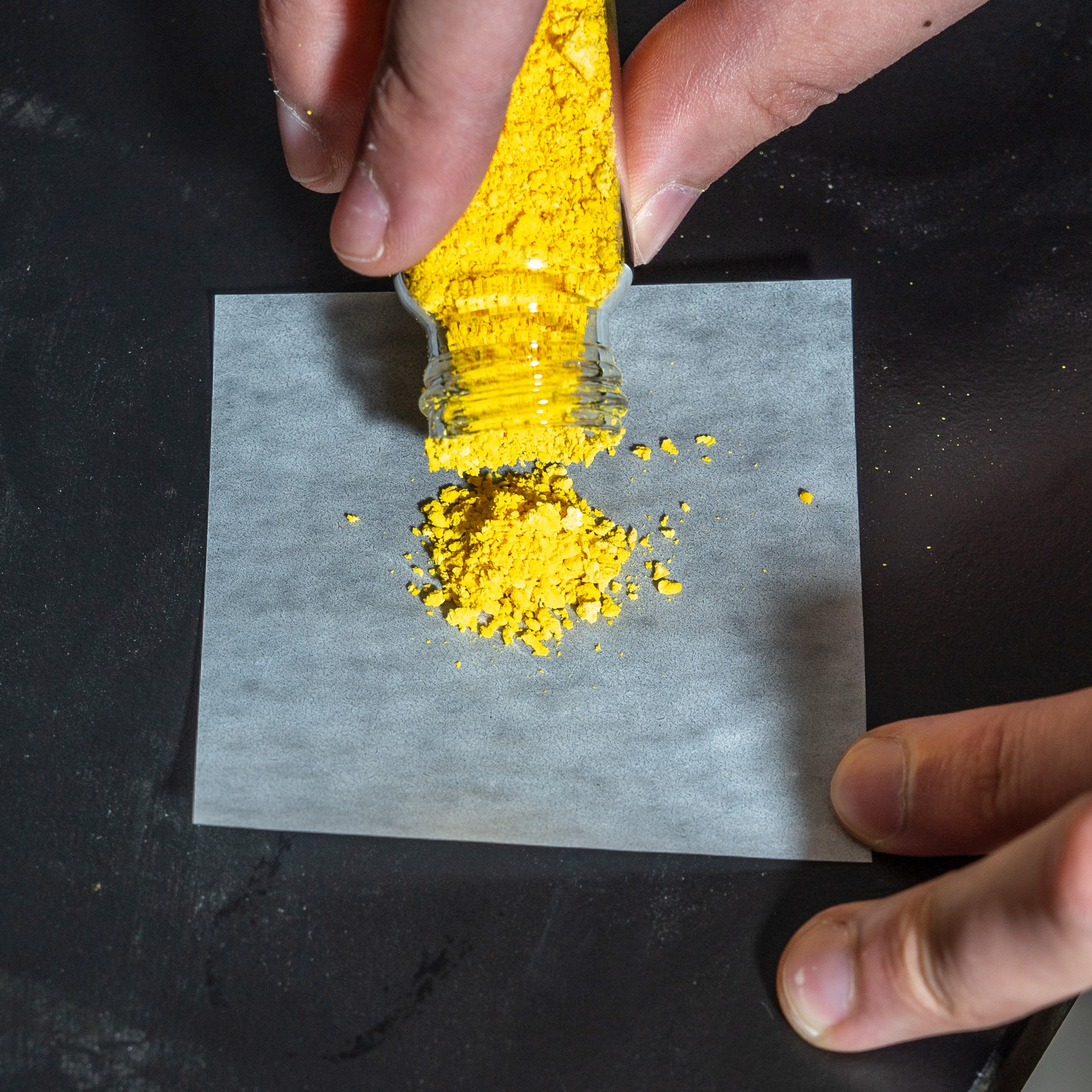 A person pours vibrant yellow powder from a small jar onto a square piece of white paper placed on a black surface.