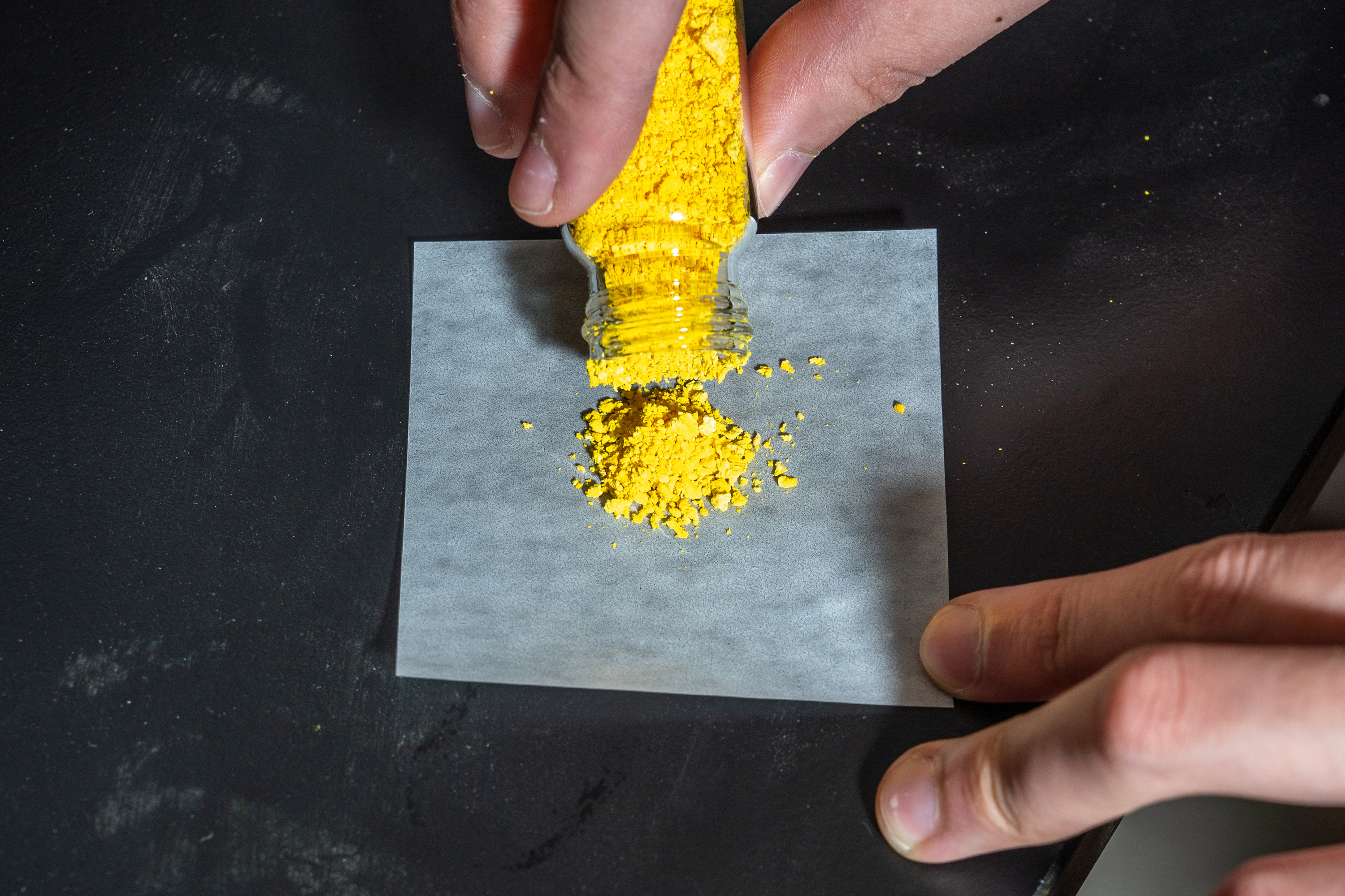 A person pours vibrant yellow powder from a small jar onto a square piece of white paper placed on a black surface.