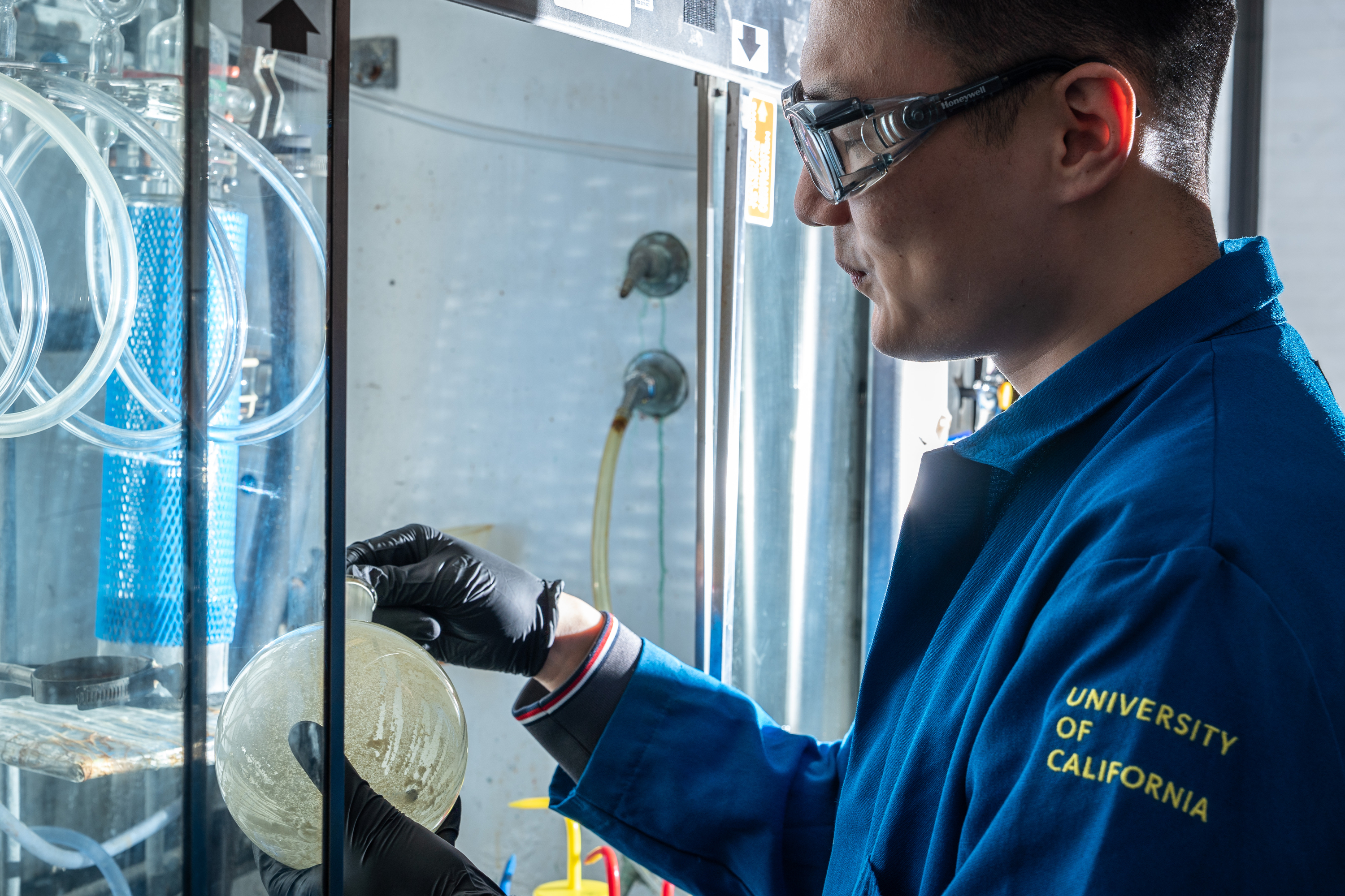 A person in a blue lab coat and safety goggles examines a round flask in a lab setting, with &quot;University of California&quot; on the coat sleeve.