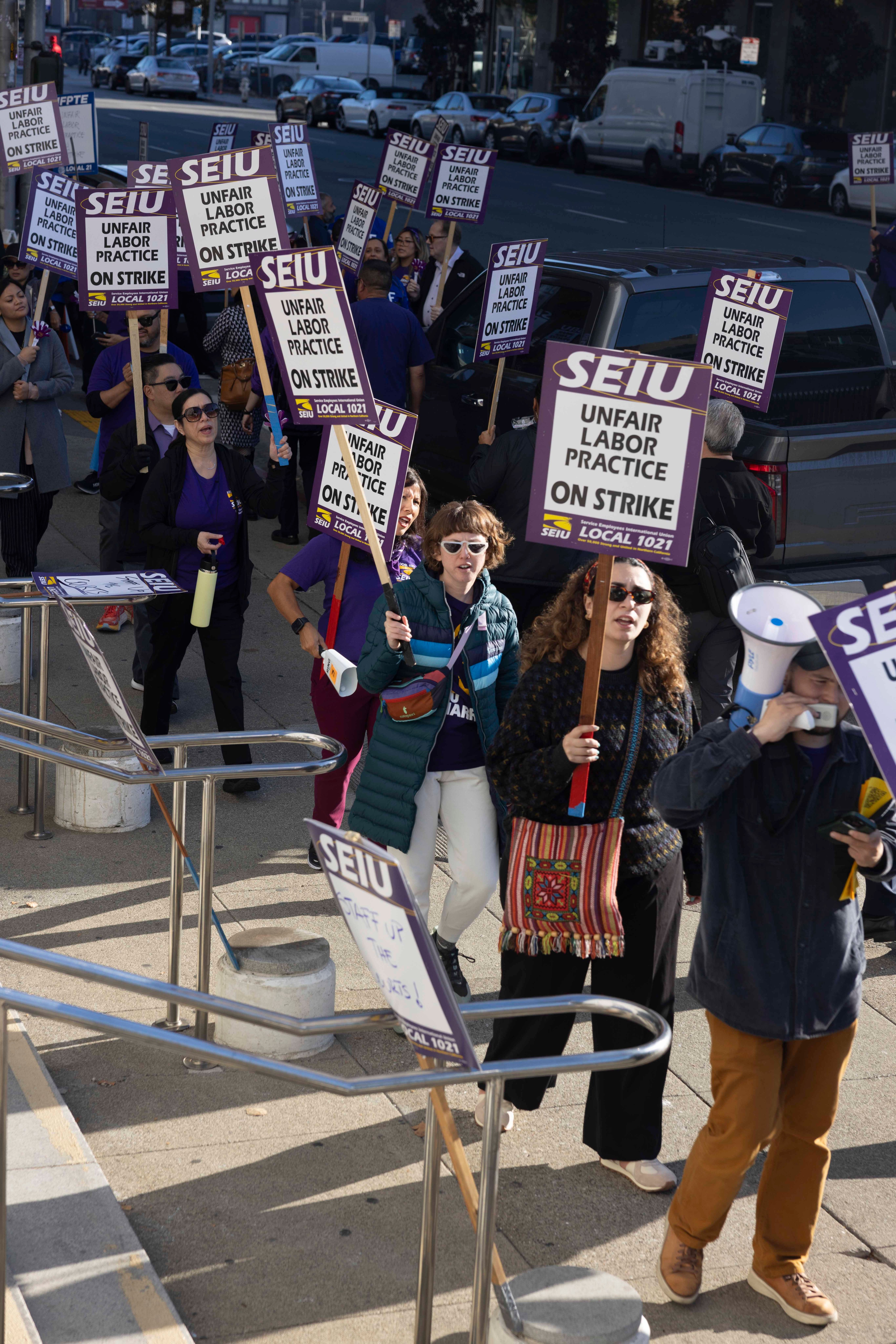 A group of protesters hold signs that read &quot;SEIU Unfair Labor Practice On Strike,&quot; marching along a sidewalk in a city, wearing sunglasses and jackets.