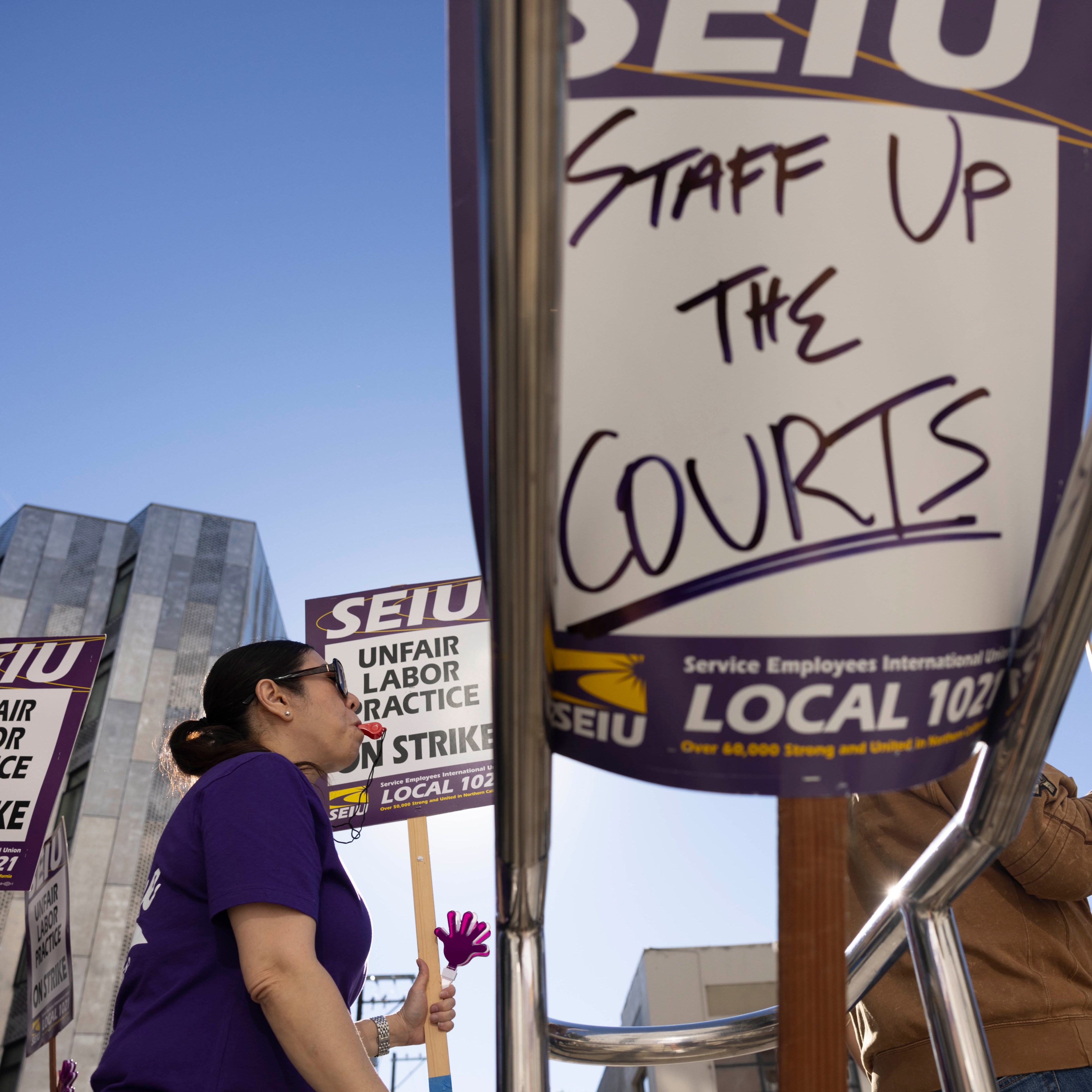 Protesters hold signs reading "SEIU Unfair Labor Practice" and "Staff Up The Courts." They stand in front of modern buildings under a clear blue sky.