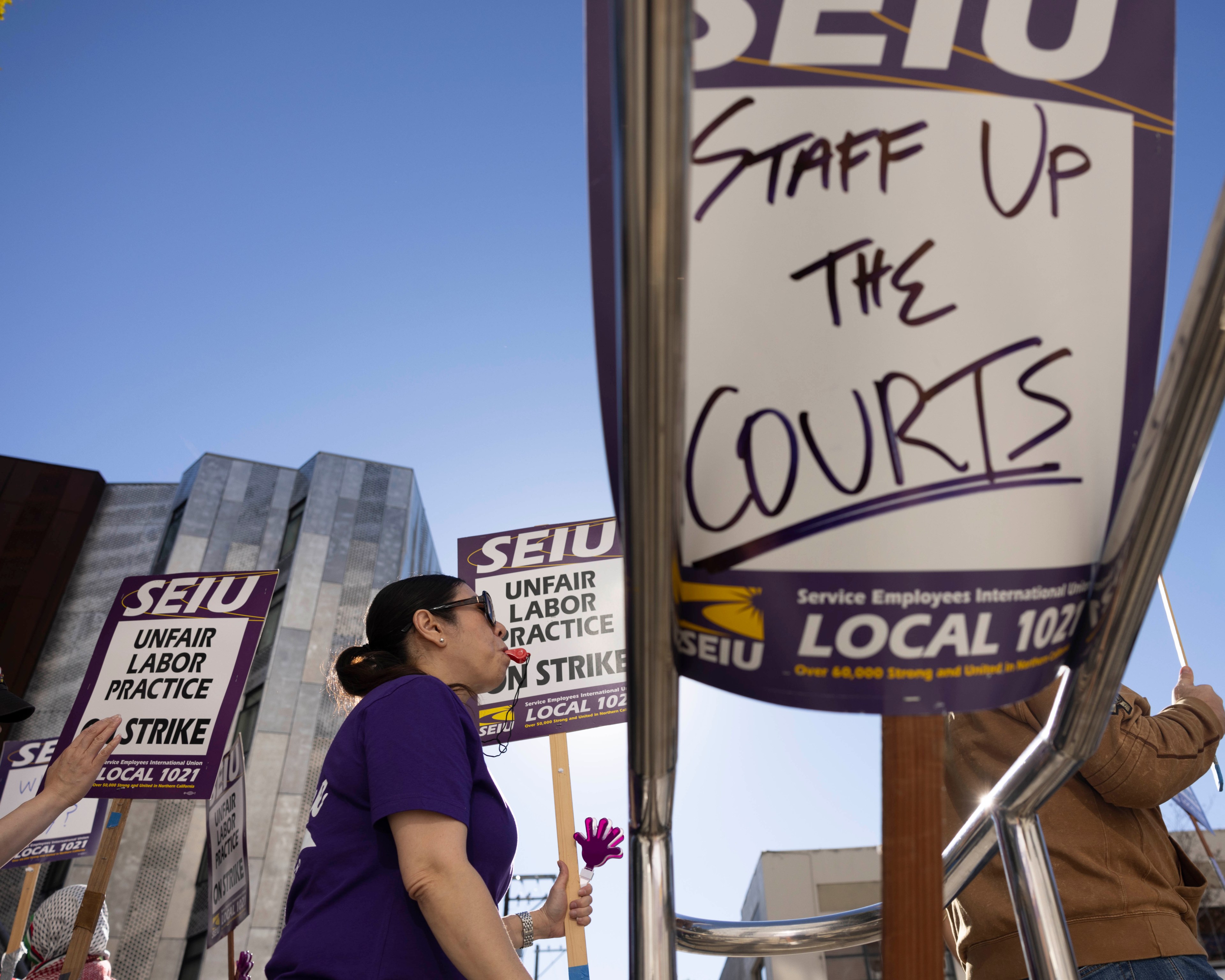 Protesters hold signs reading "SEIU Unfair Labor Practice" and "Staff Up The Courts." They stand in front of modern buildings under a clear blue sky.