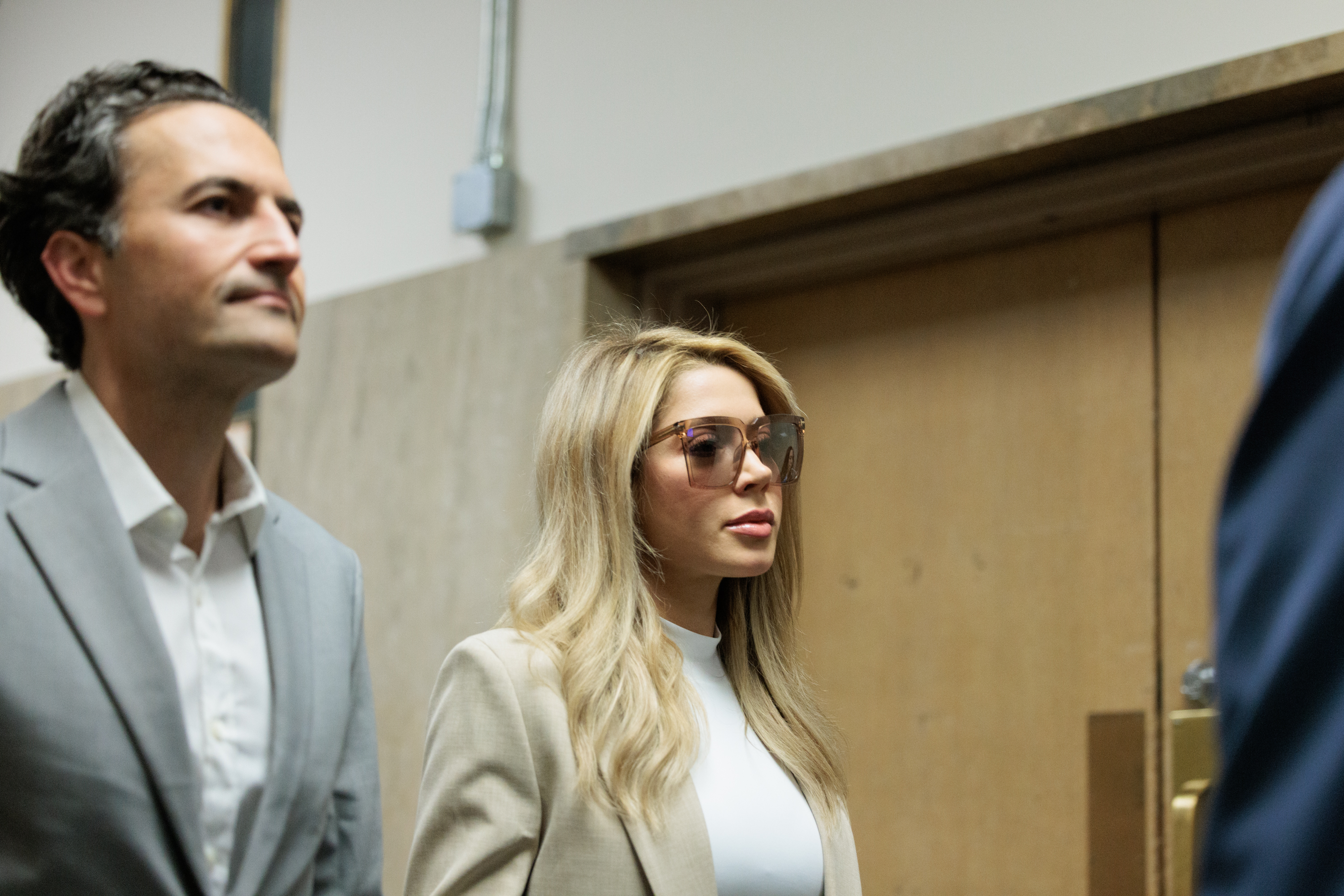 A blonde woman in sunglasses and a light-colored suit walks alongside a man in a gray suit. They appear to be in a hallway with wooden doors.