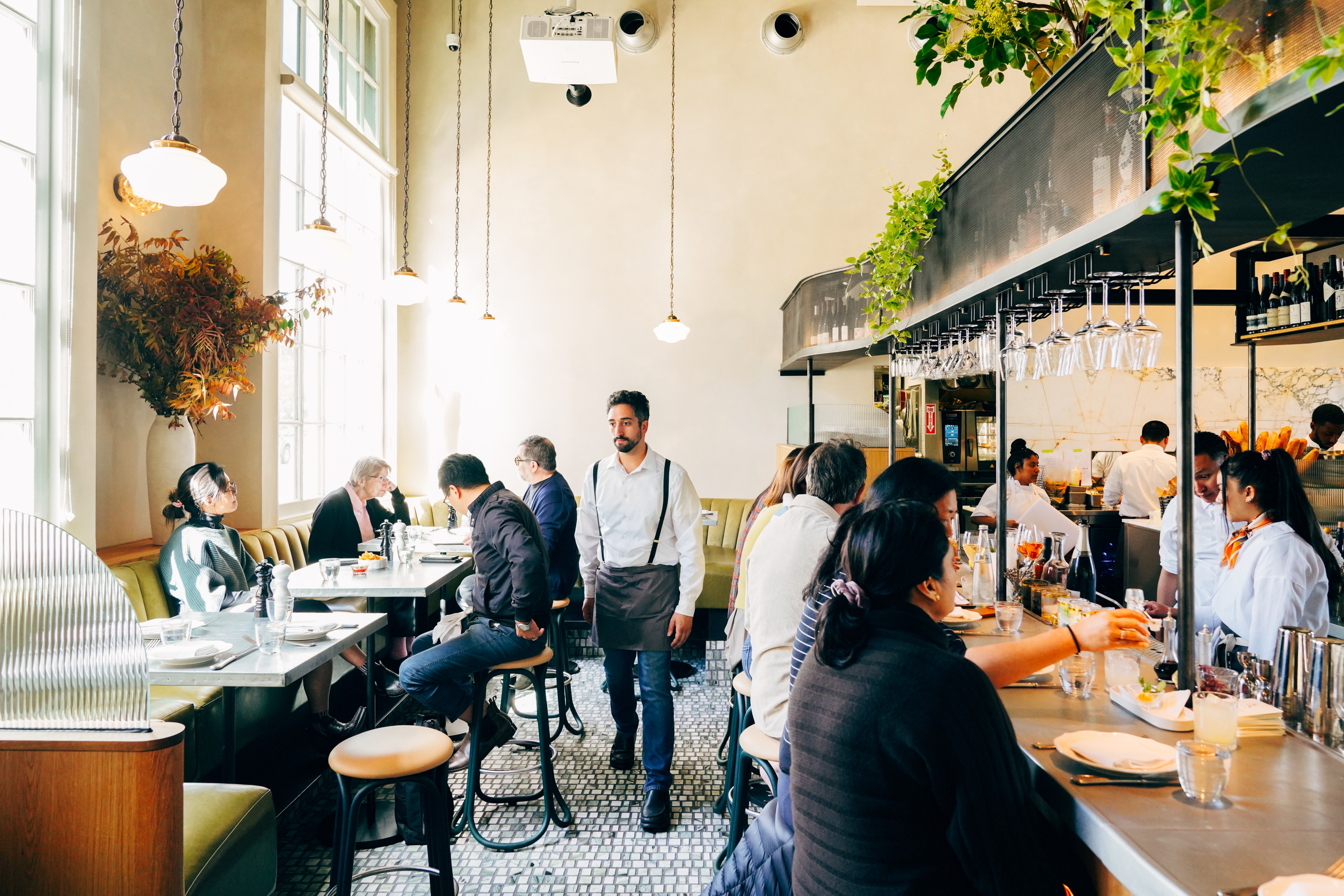 People are dining in a bright, modern restaurant with pendant lights, greenery, and large windows. A waiter walks through the room, and bartenders serve drinks.