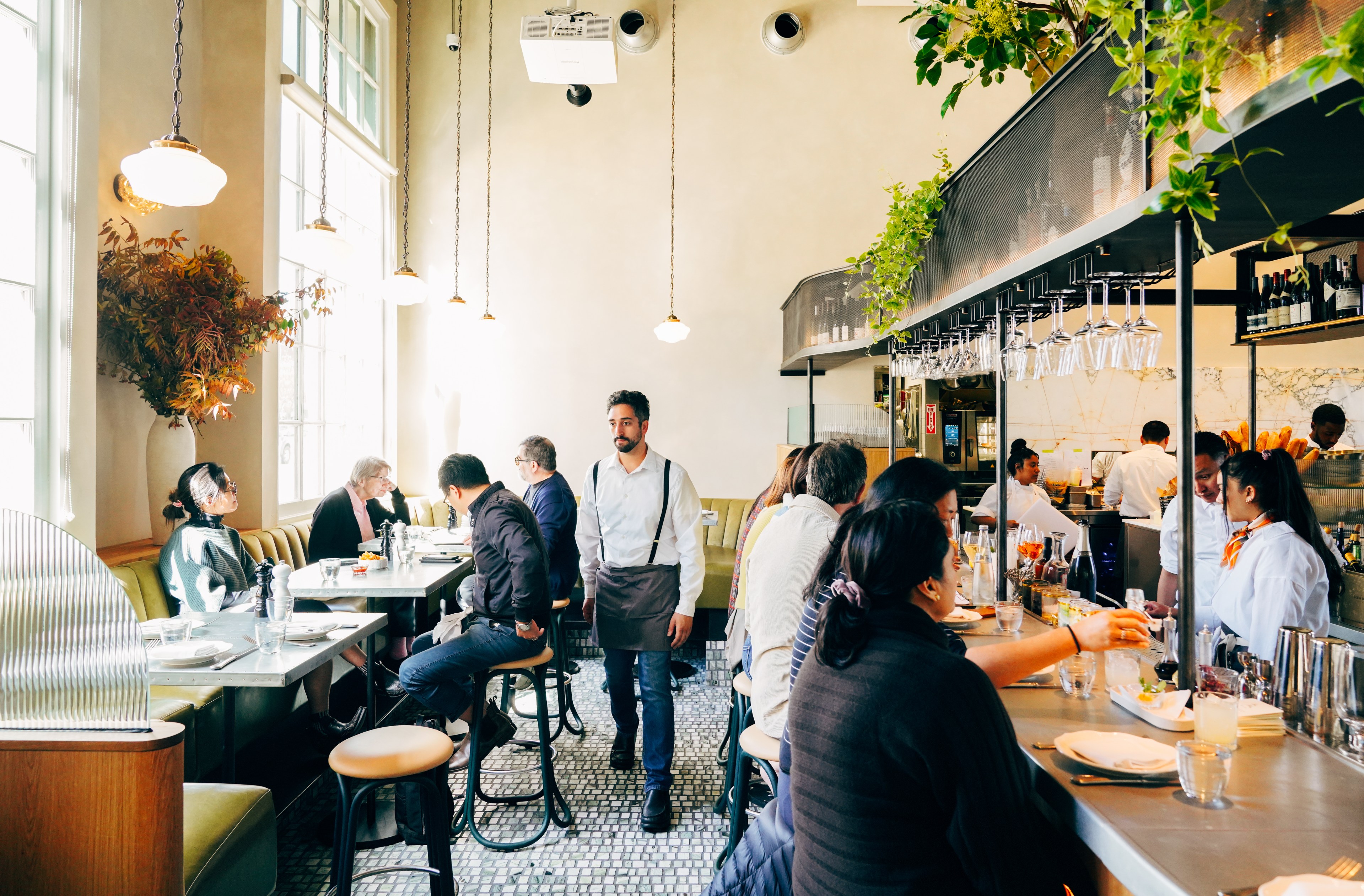 People are dining in a bright, modern restaurant with pendant lights, greenery, and large windows. A waiter walks through the room, and bartenders serve drinks.