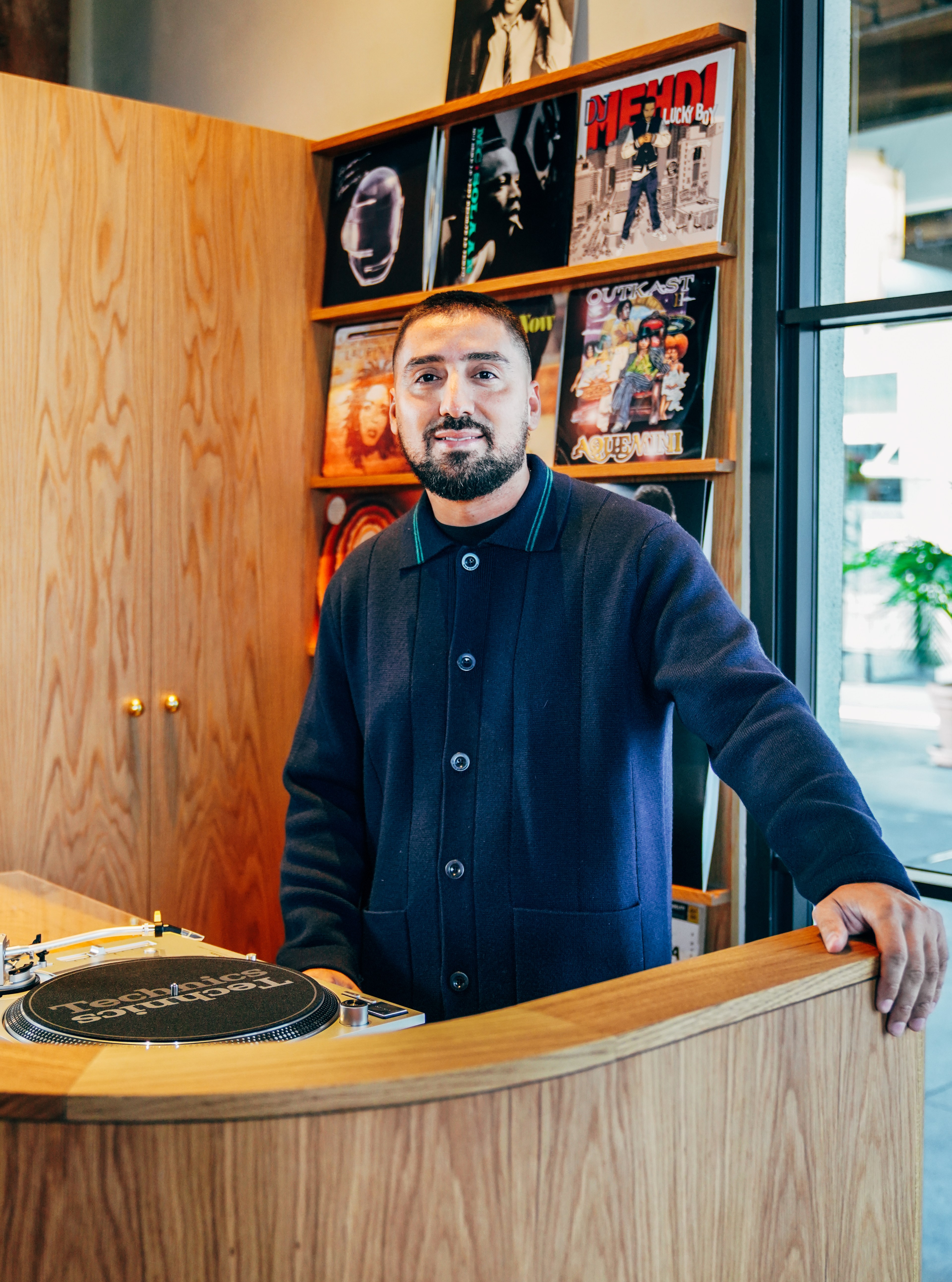 A man stands behind a wooden DJ booth with a turntable. In the background, there are shelves displaying various music records.