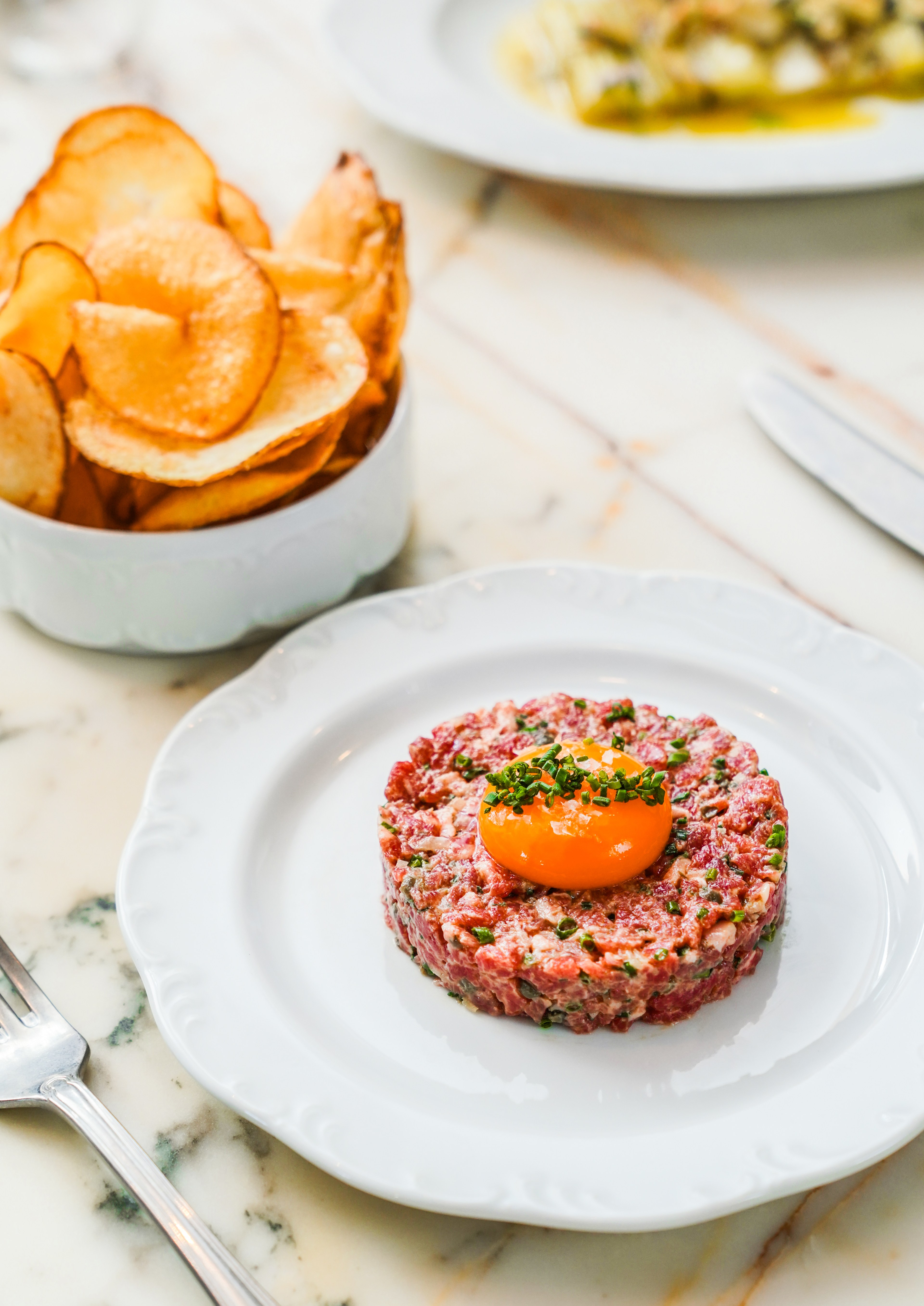 A plate of steak tartare topped with an egg yolk and herbs sits on a marble table. Next to it is a bowl of thinly sliced, crispy chips.