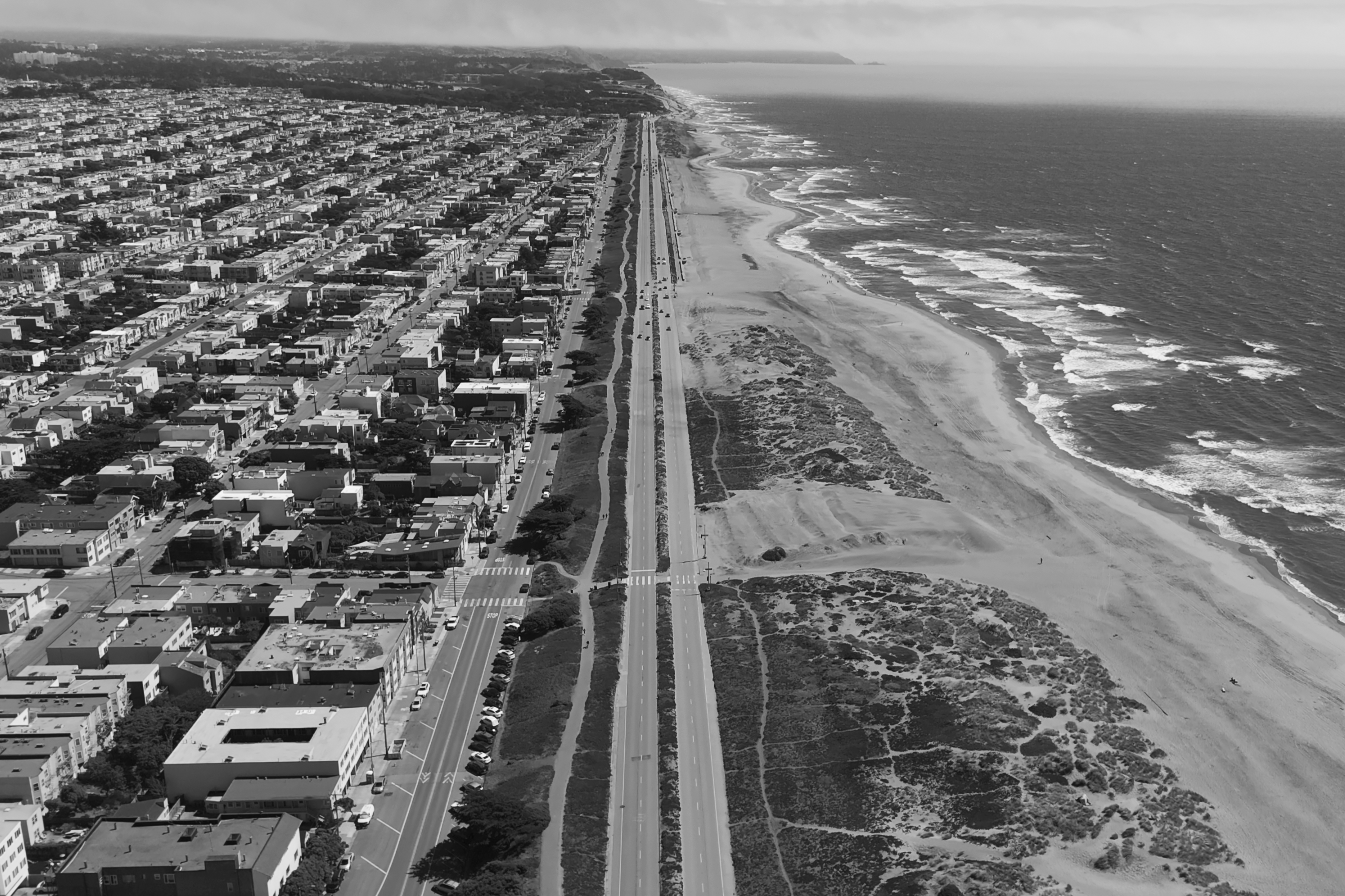 The image shows a coastal cityscape with a long stretch of beach to the right and densely packed buildings to the left, divided by a road running parallel to the shoreline.