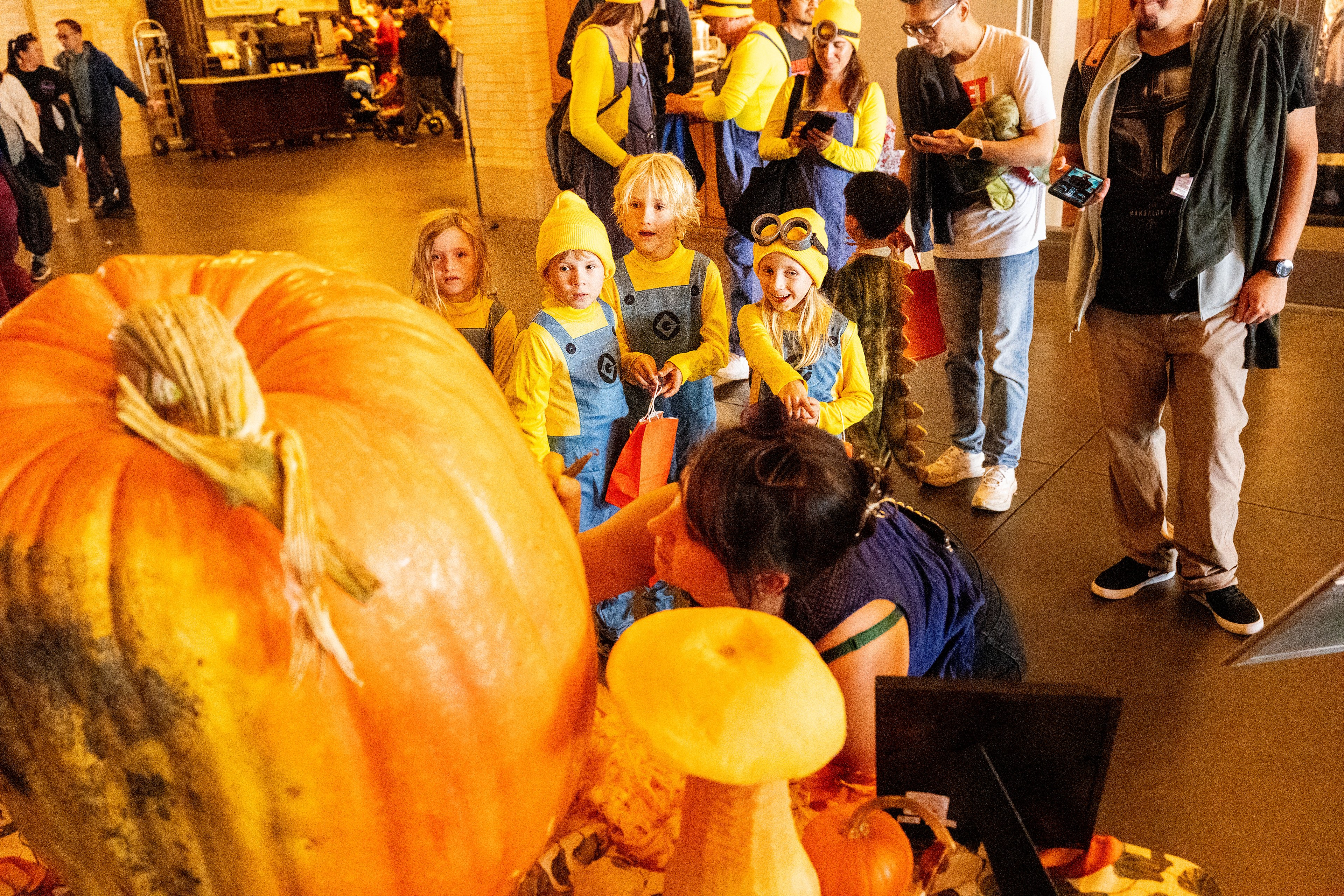 Children in Minion costumes gather around a large pumpkin. An adult is with them, some people in the background, and various pumpkins and decorations are visible.