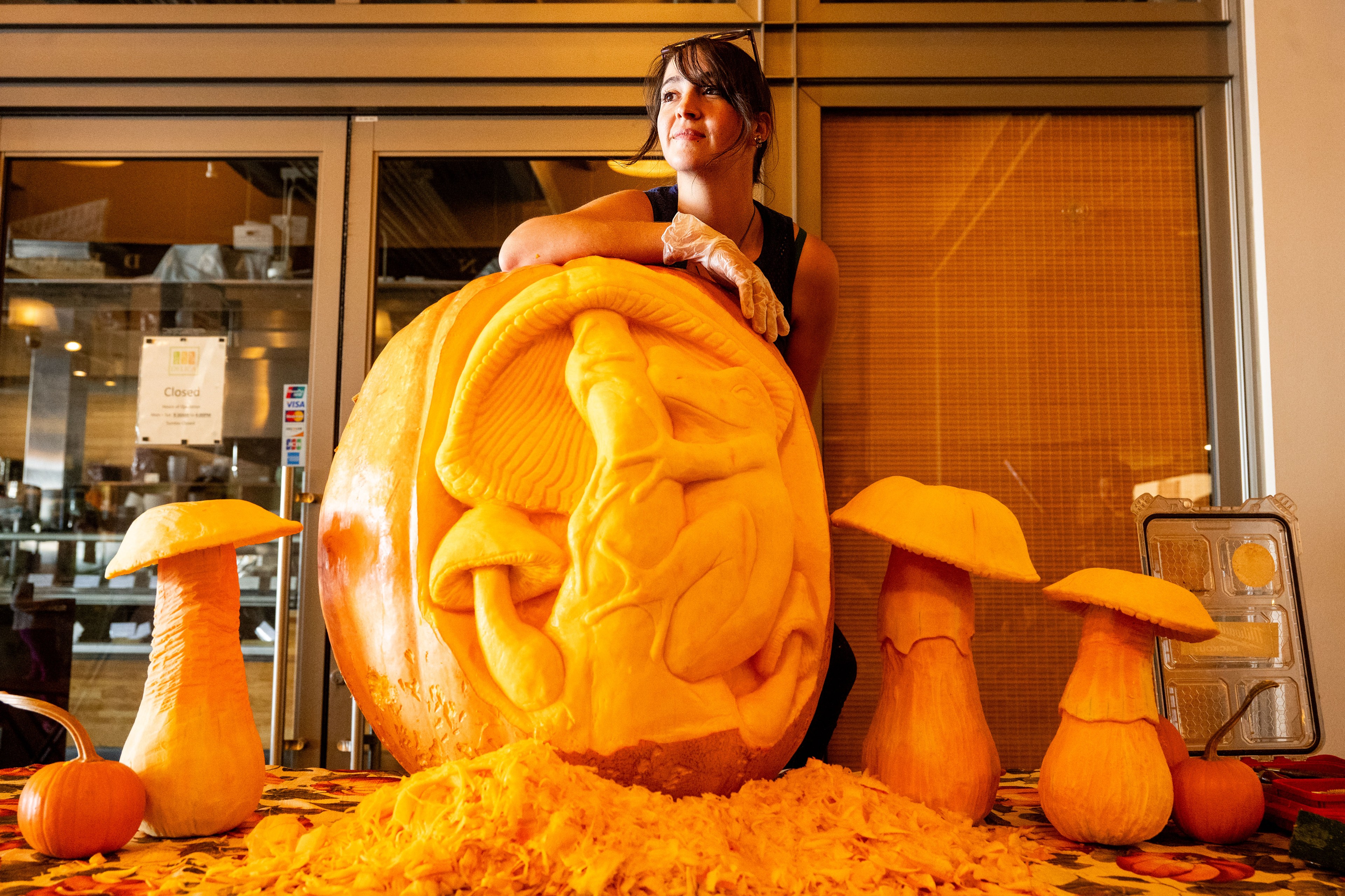 A person leans on a large intricately carved pumpkin featuring a detailed frog and mushroom design, surrounded by pumpkin shavings and smaller carved mushrooms.