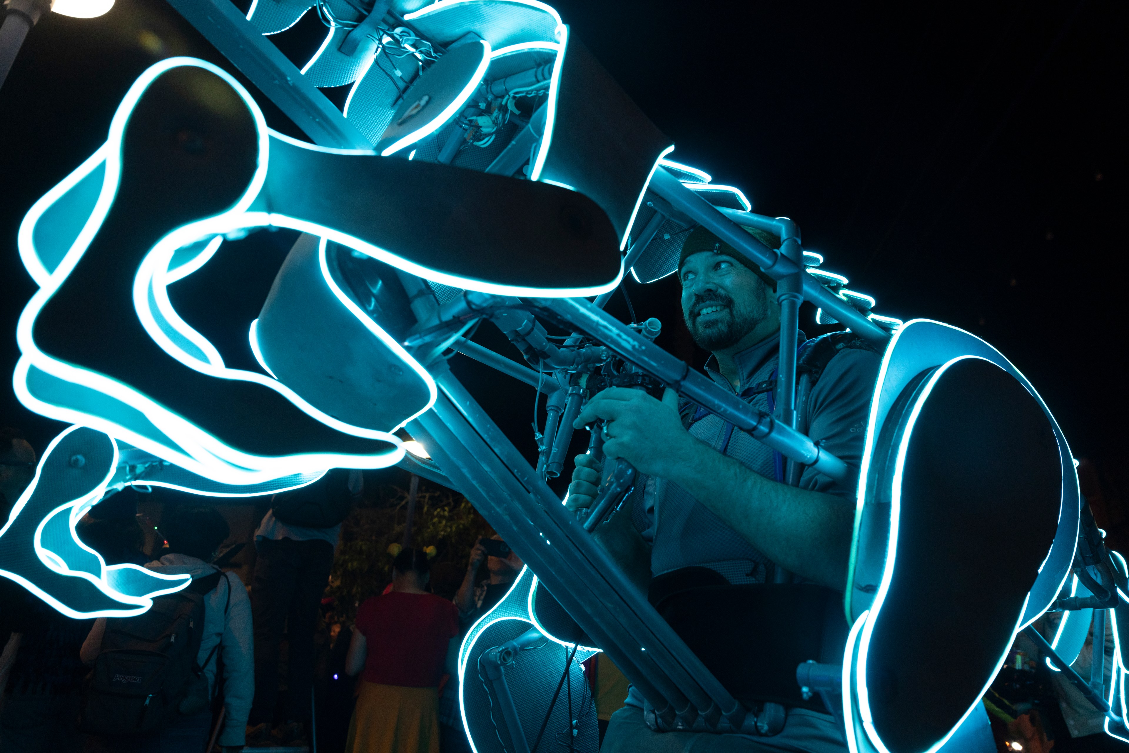 A person smiles while playing an instrument within a glowing, neon-lit structure at night. The abstract design creates a vibrant, futuristic feel.