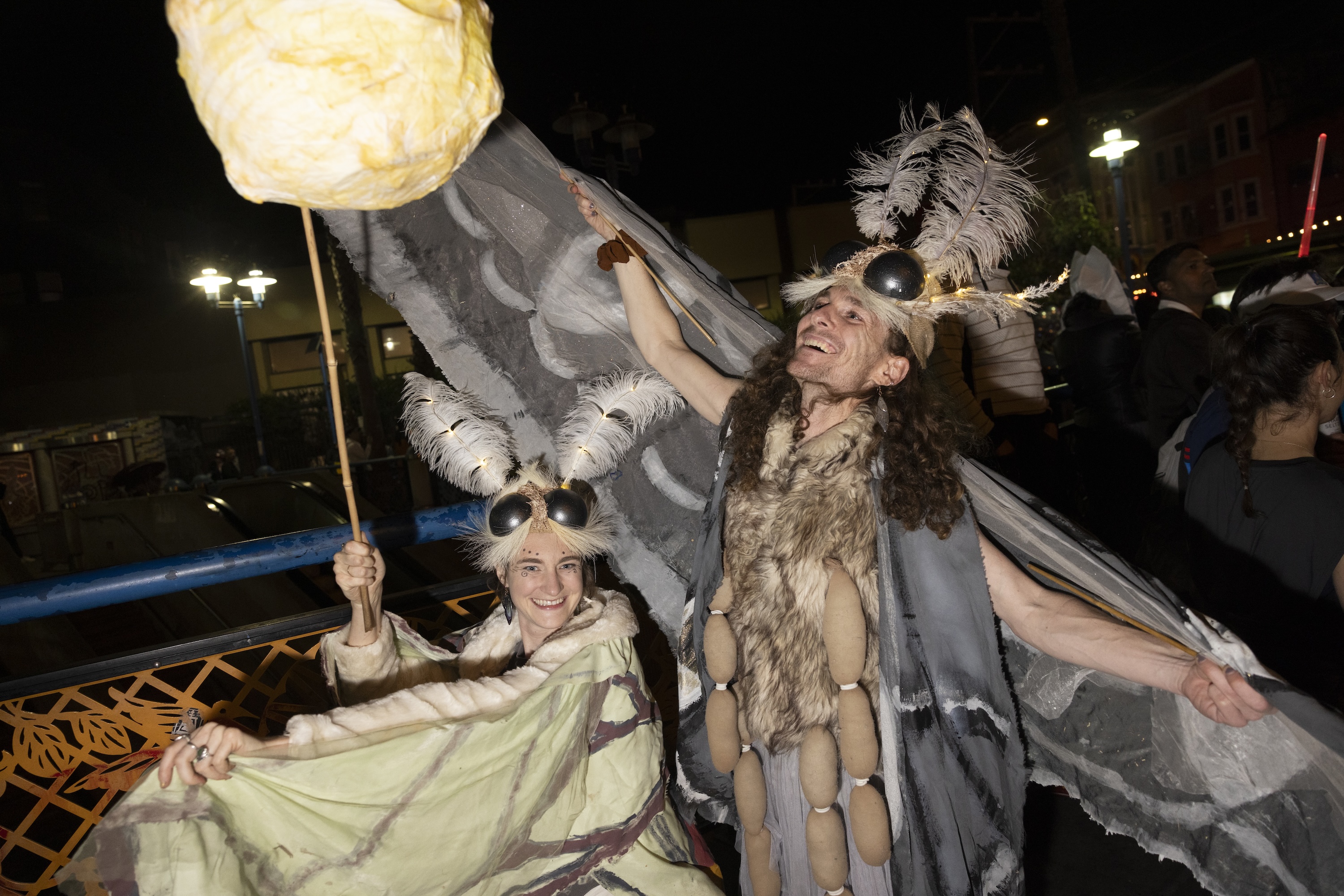 Two people joyfully dressed as moths, complete with feathered antennae and large wings, hold a large glowing orb against a dimly lit urban background.