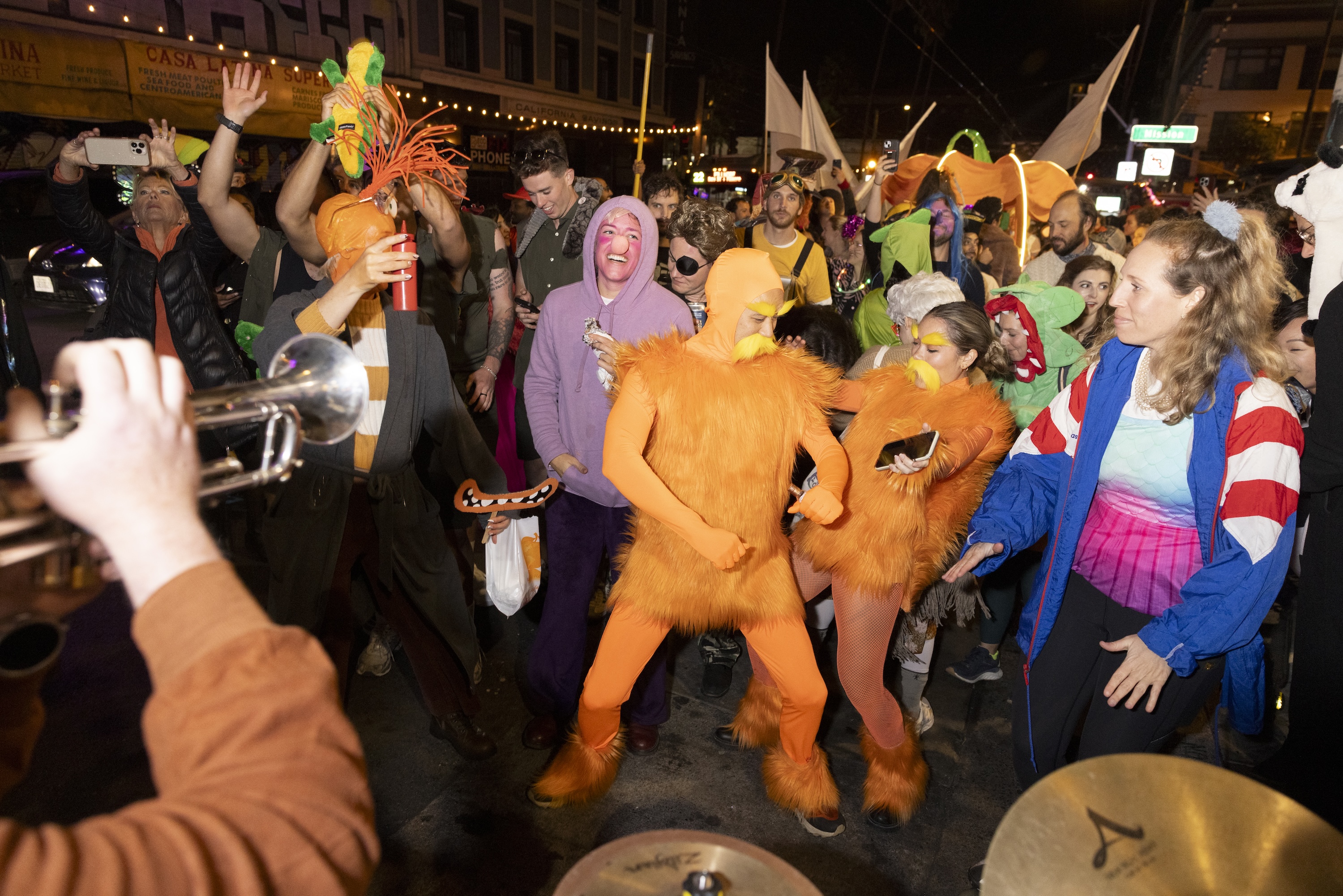 A lively crowd of people in colorful, eccentric costumes are dancing energetically at an outdoor event, with a musician playing a trumpet in the foreground.