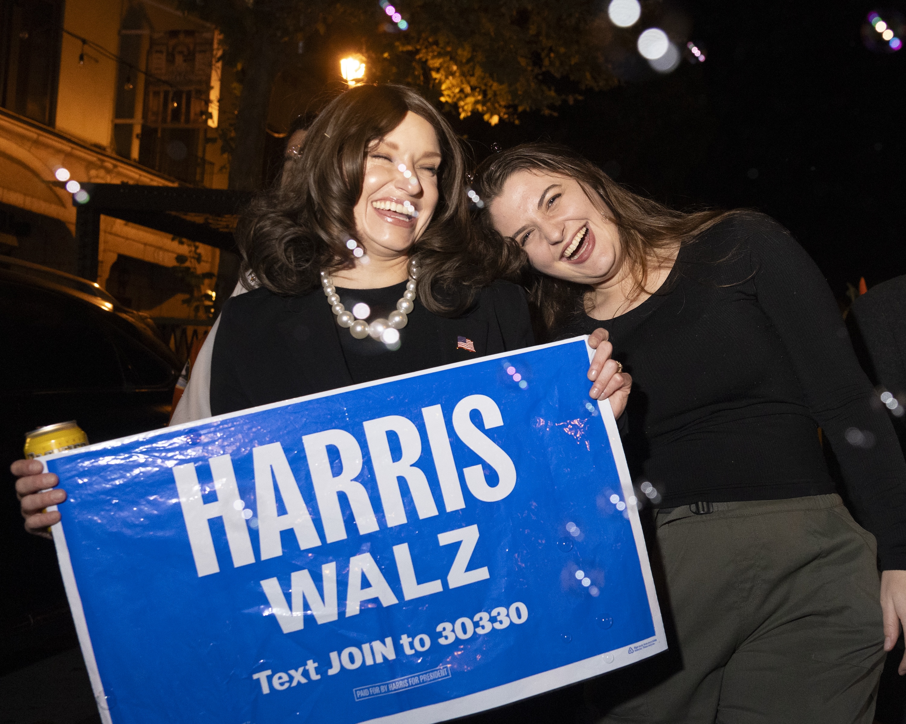 Two people are smiling and holding a blue sign that reads &quot;HARRIS WALZ&quot; with a message to text JOIN to a number. One person is holding a drink can.