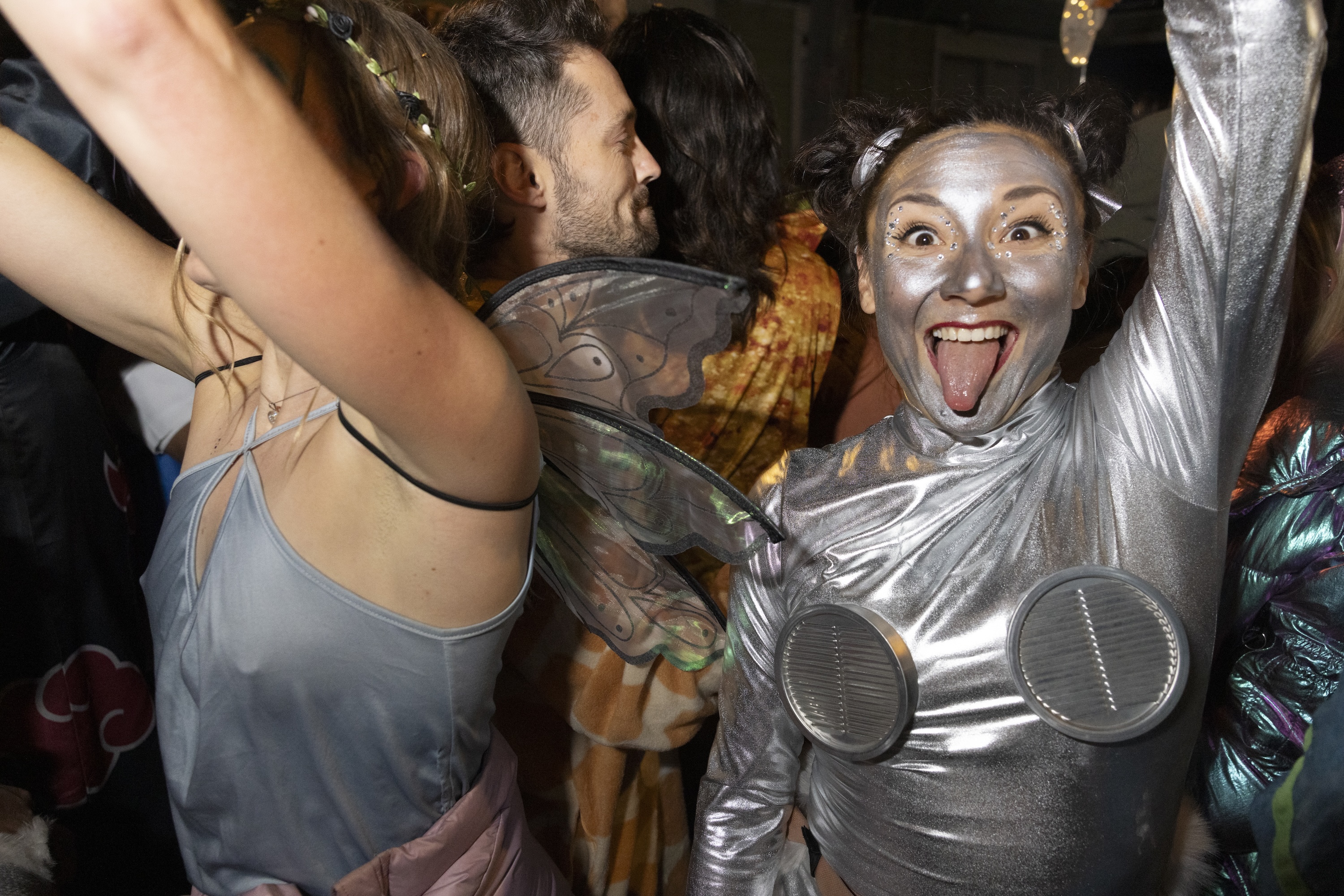 A lively party scene with a woman in shiny silver face paint and costume, grinning and sticking out her tongue. People around her wear colorful, whimsical outfits.