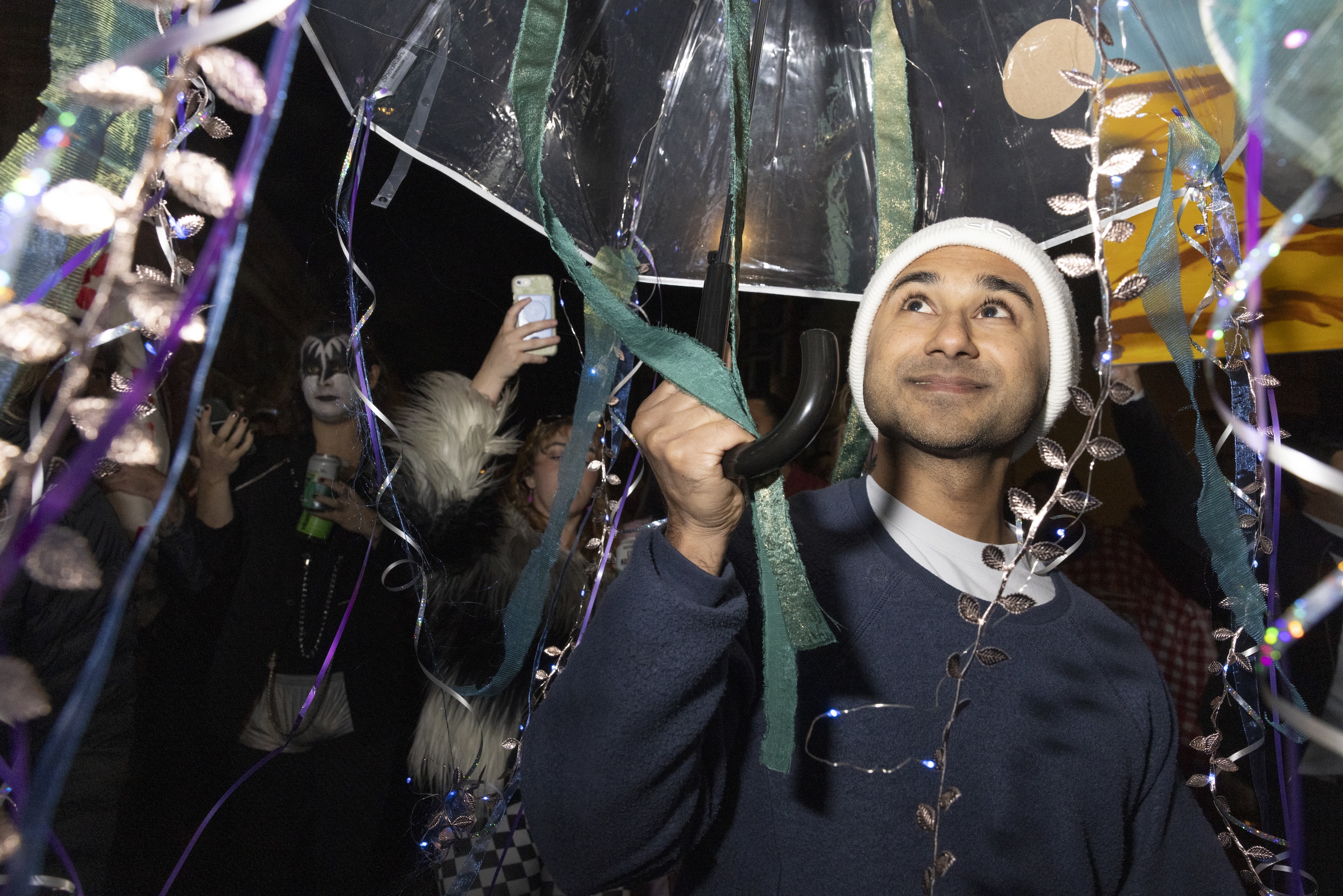 A person in a beanie smiles under a decorated umbrella with ribbons and lights, surrounded by people, one of whom is wearing face paint and a costume.