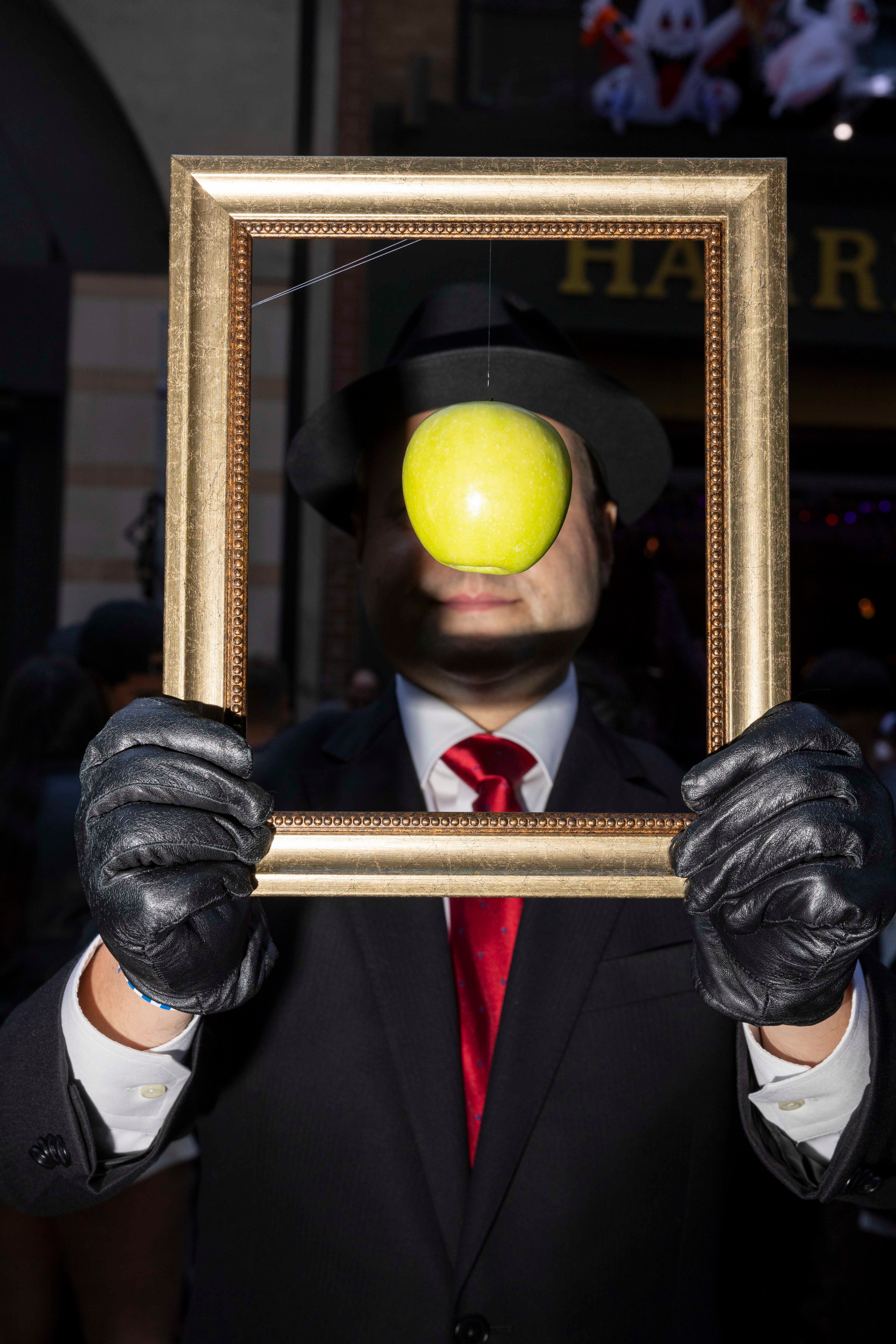 A person in a suit and hat is holding a golden frame with gloved hands. A green apple hangs in front of their face, obscuring their features.