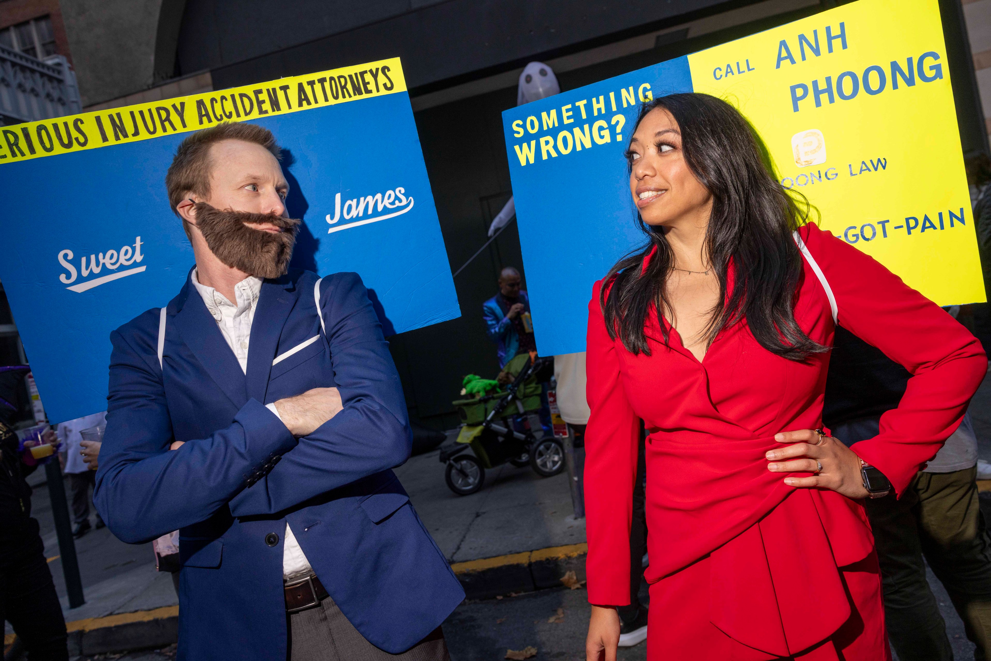 Two people in costumes stand facing each other with blue and yellow signs behind them, dressed as lawyers. One wears a blue suit, the other a red outfit.