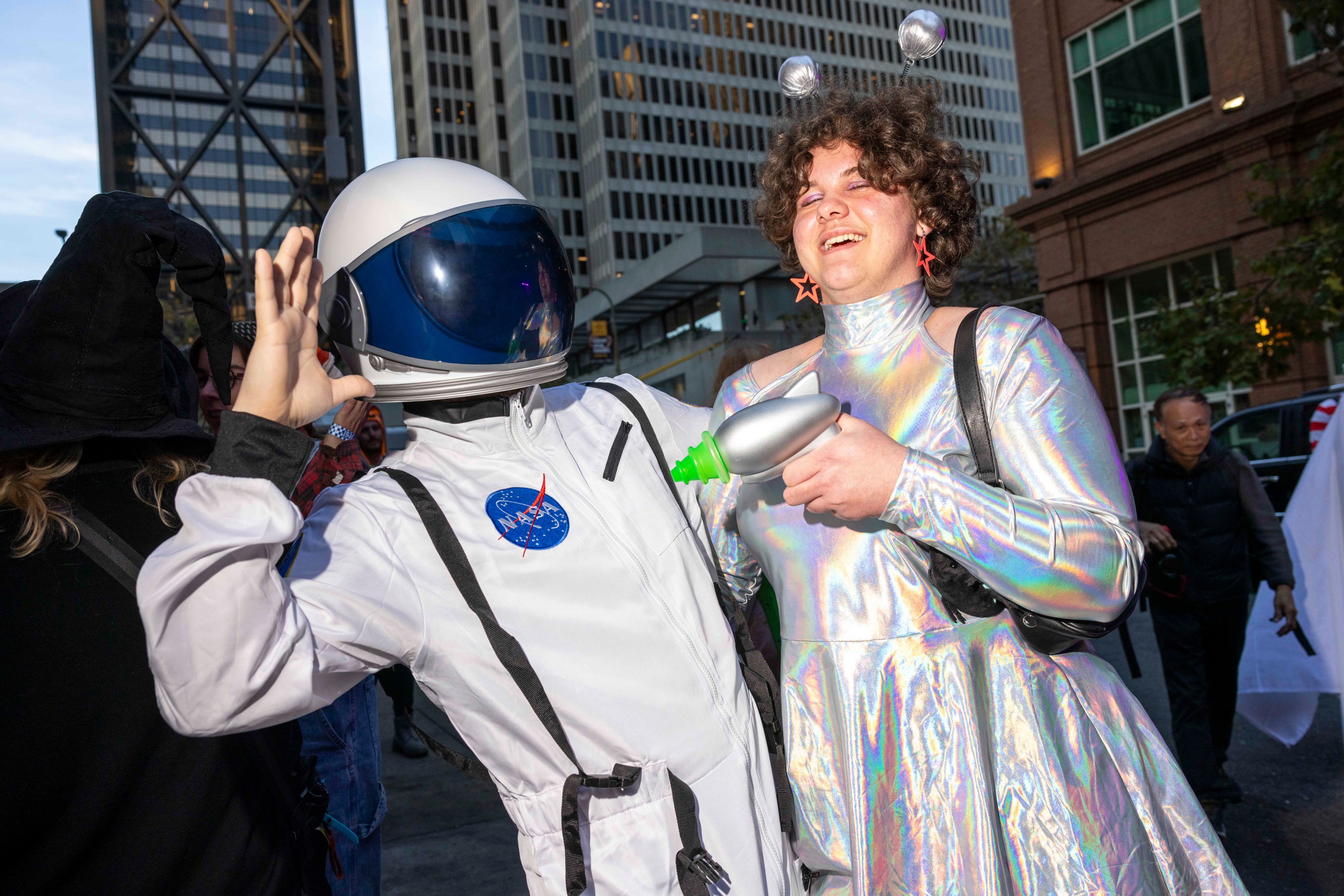 Two people are in costume on a city street. One wears a NASA astronaut suit, and the other is in a shiny alien outfit with antennae and a toy ray gun.