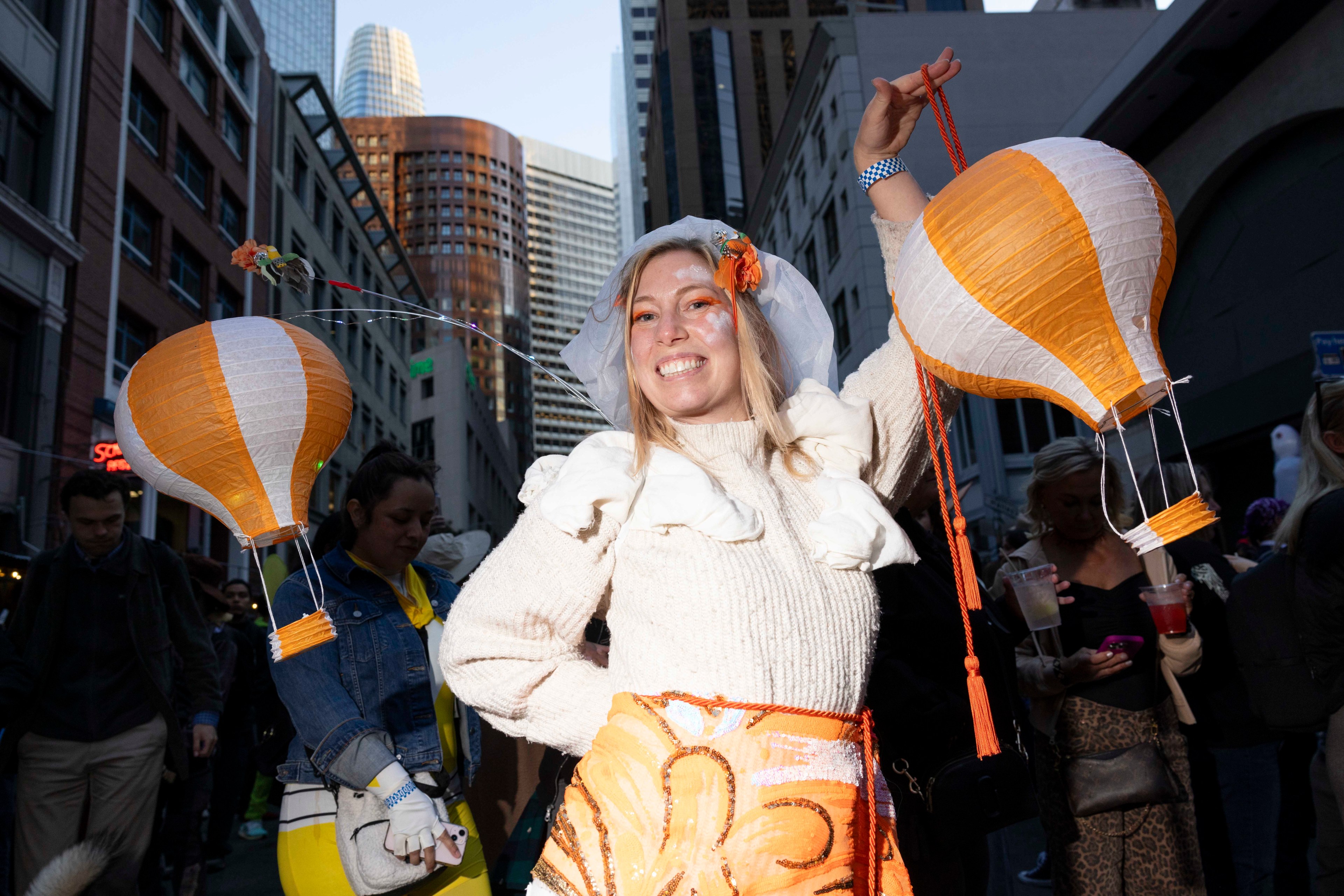 A smiling person in an urban setting holds two orange and white balloon-shaped lanterns. They wear a white top with decorative elements and are surrounded by people.