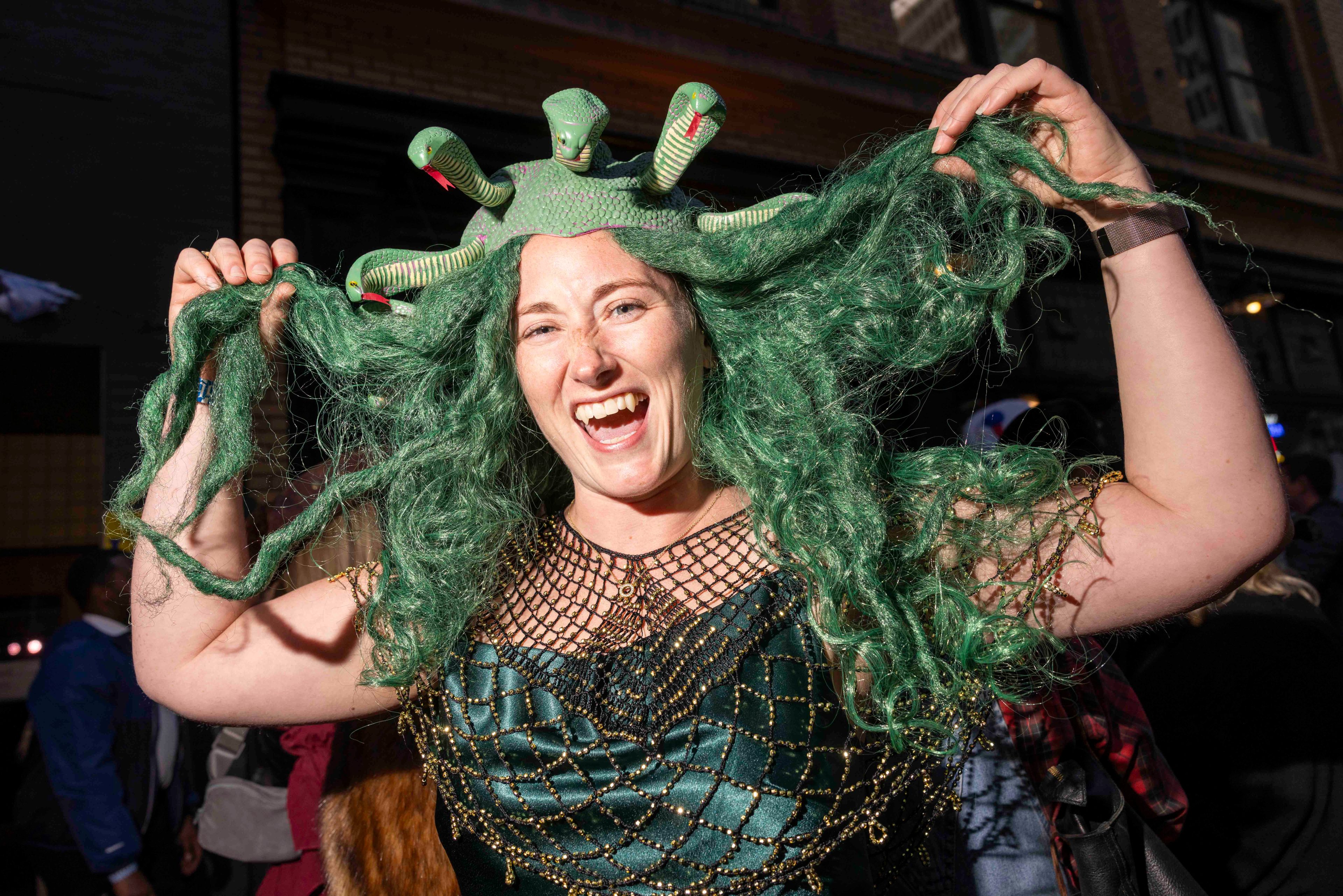 A person smiles widely while wearing a Medusa costume with green snake-like hair and a detailed green dress. They are holding their hair up playfully.