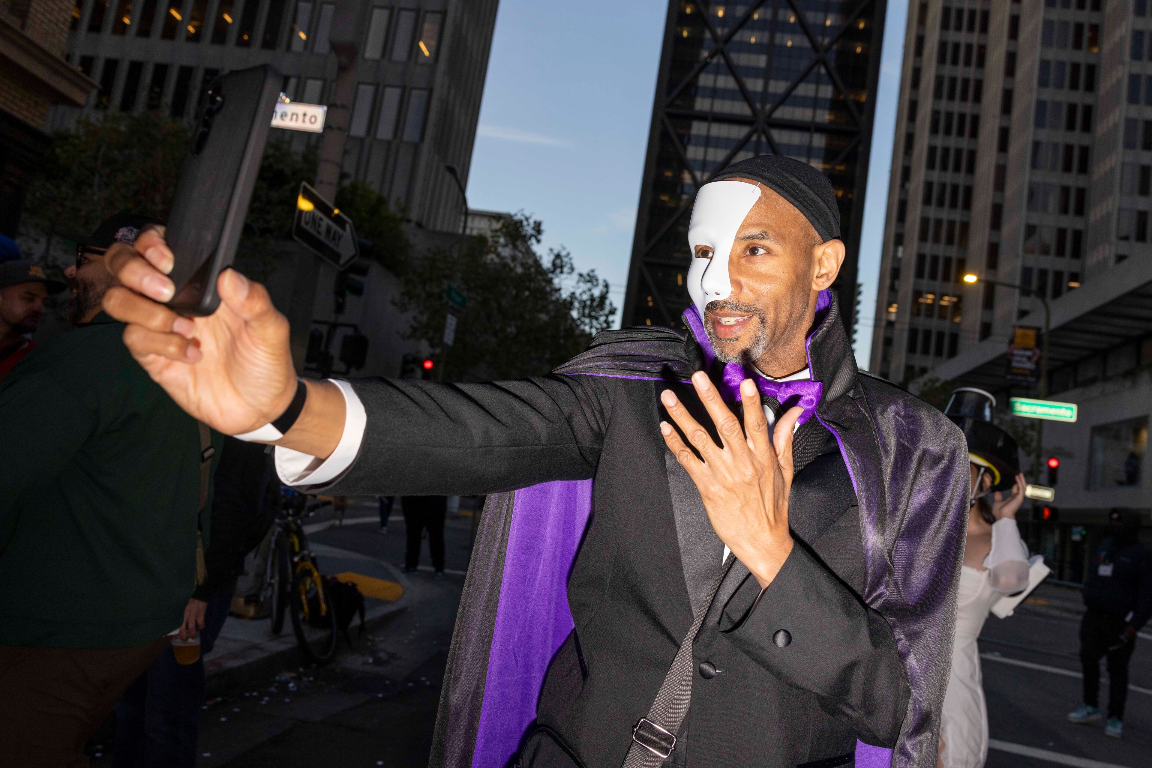 A man in a half-face white mask and black suit takes a selfie in a city street, wearing a purple cape and bow tie, with tall buildings in the background.