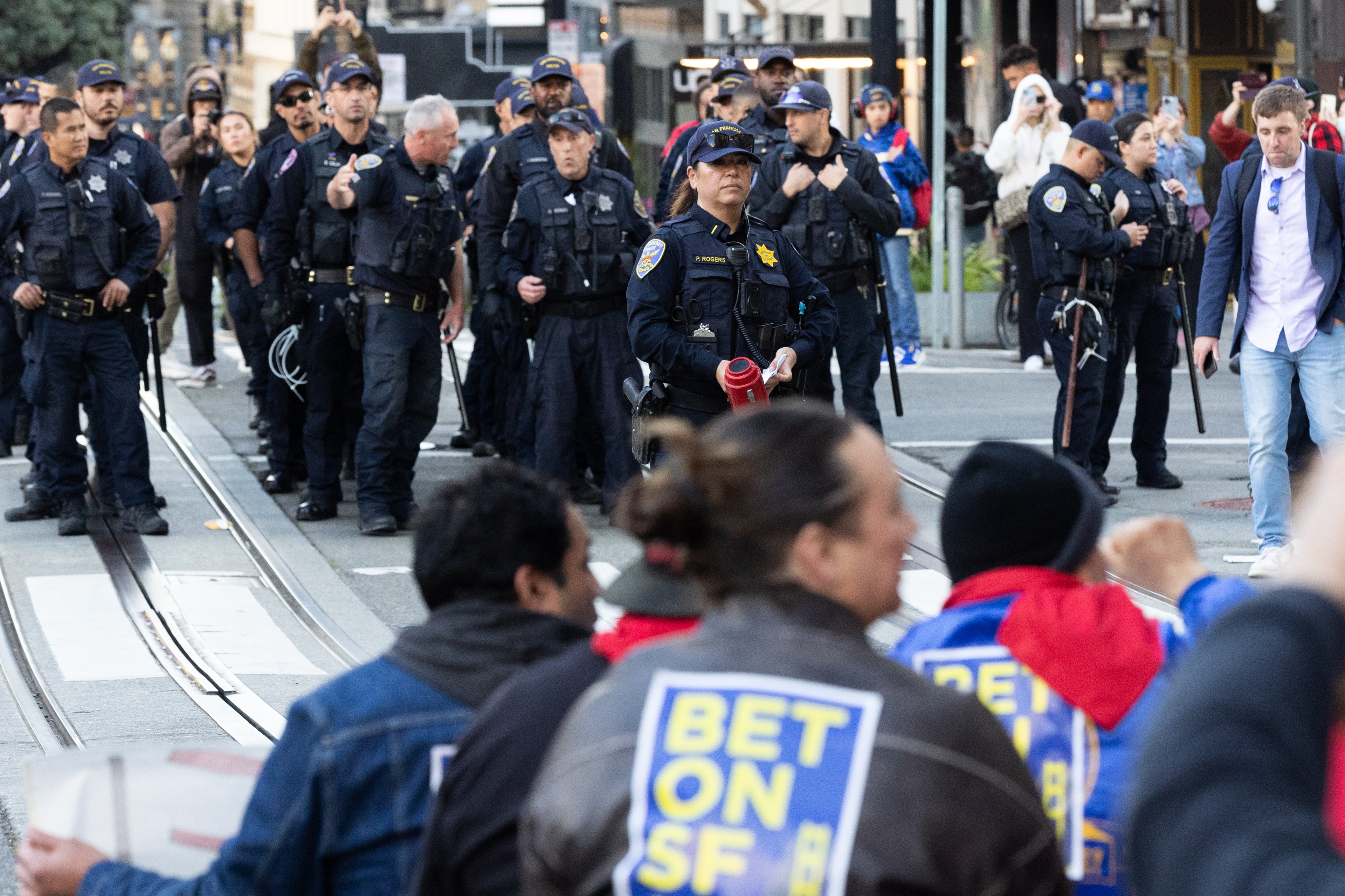 A group of police officers stand on a street facing a crowd of seated protestors. The protestors wear signs with &quot;BET ON SF&quot; on their backs.