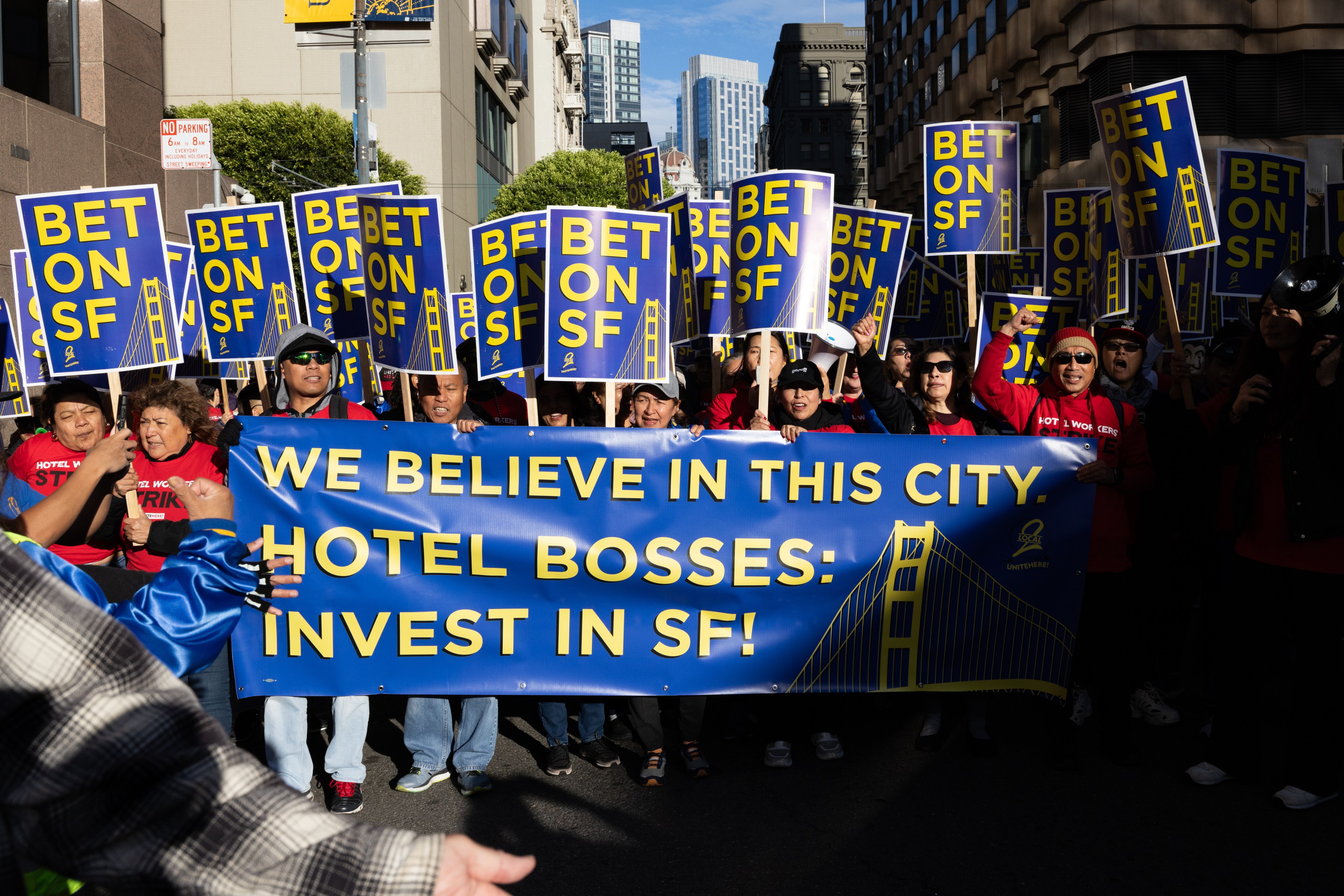 A group of people are marching on a city street, holding signs that read &quot;BET ON SF&quot; and a large banner that says &quot;WE BELIEVE IN THIS CITY. HOTEL BOSSES: INVEST IN SF!&quot;