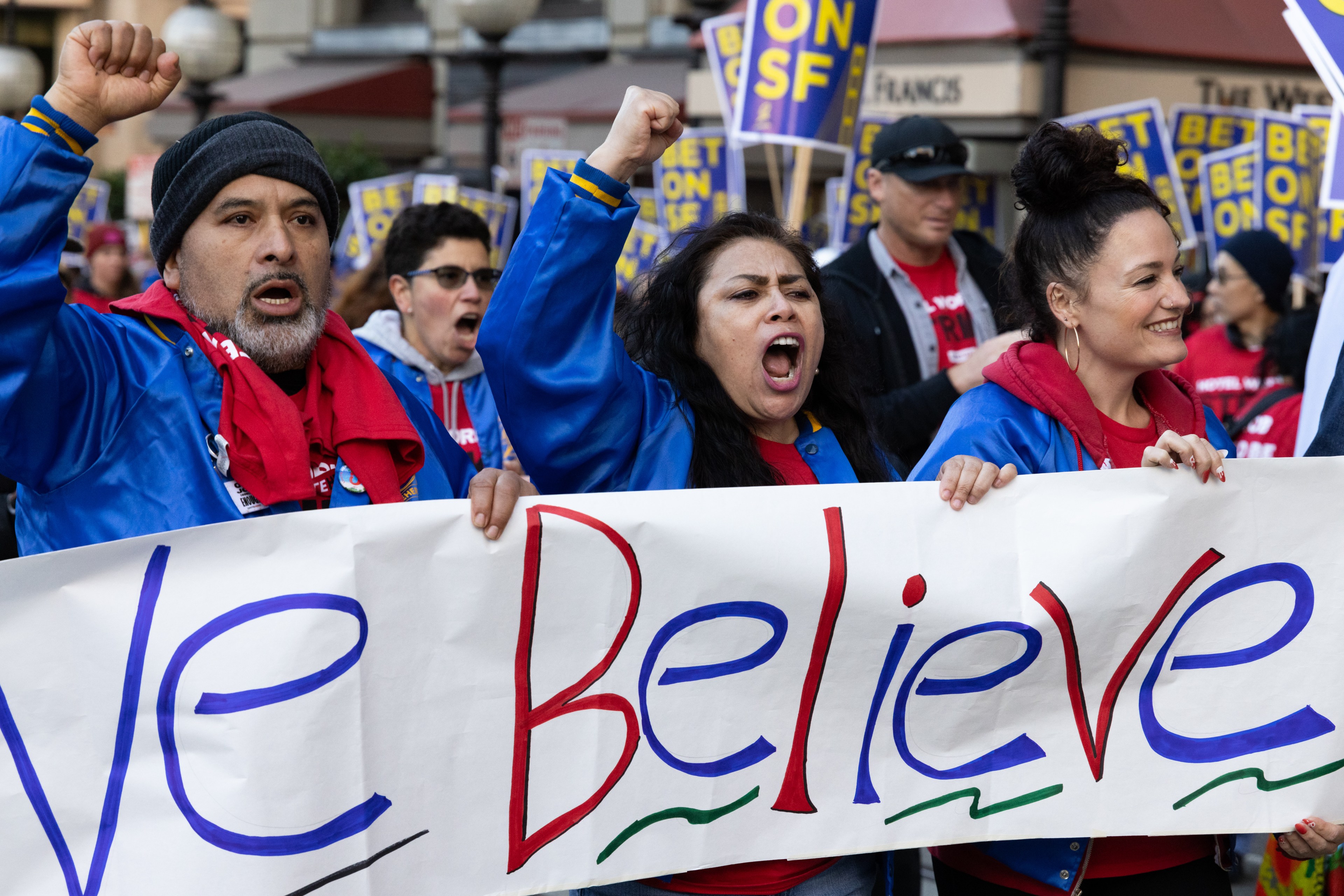 A group of people in blue jackets and red shirts hold up a sign reading &quot;Believe&quot; while shouting and raising their fists during a protest or rally.