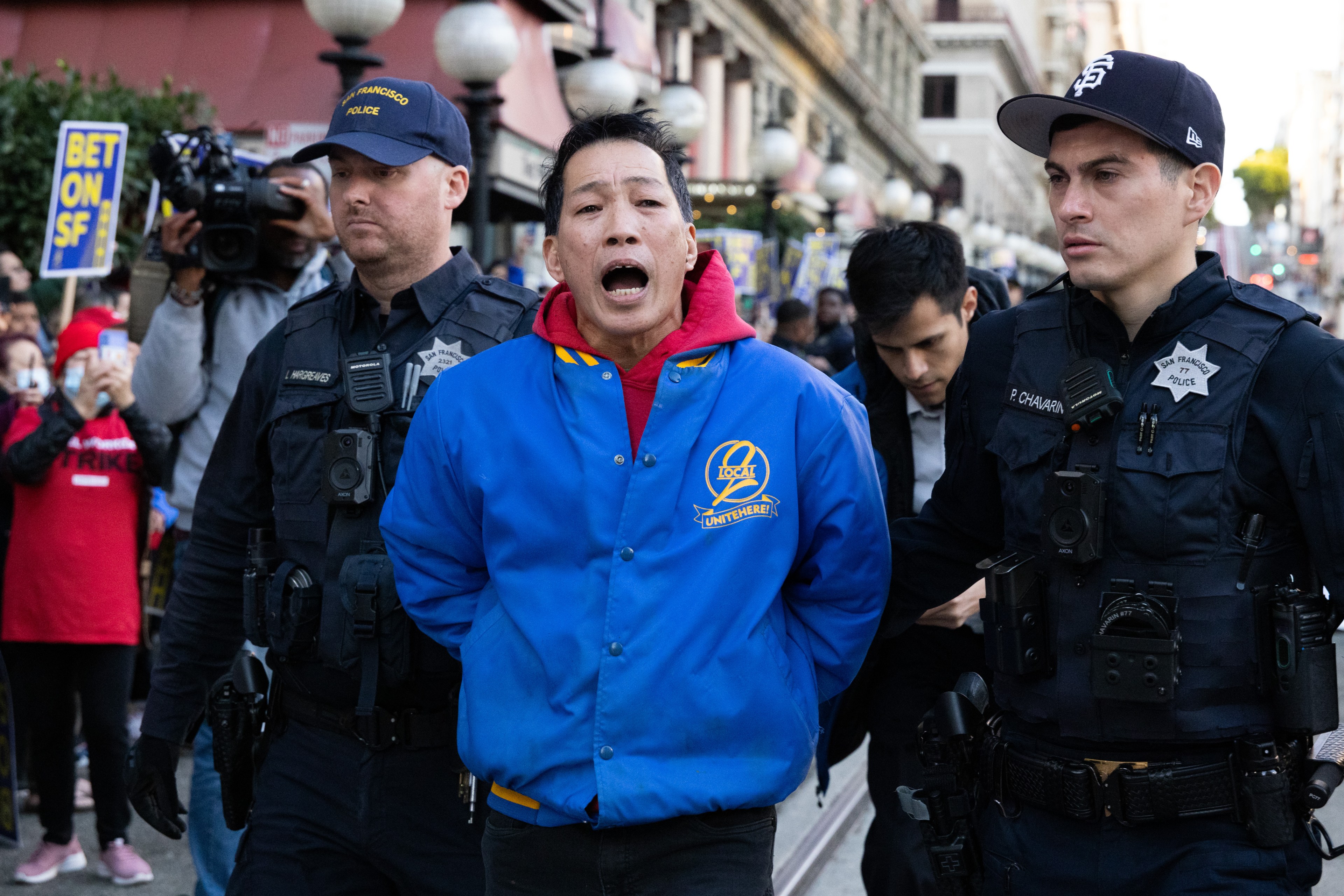 A man in a blue jacket is being escorted by two police officers in dark uniforms. There is a crowd in the background, some holding signs.