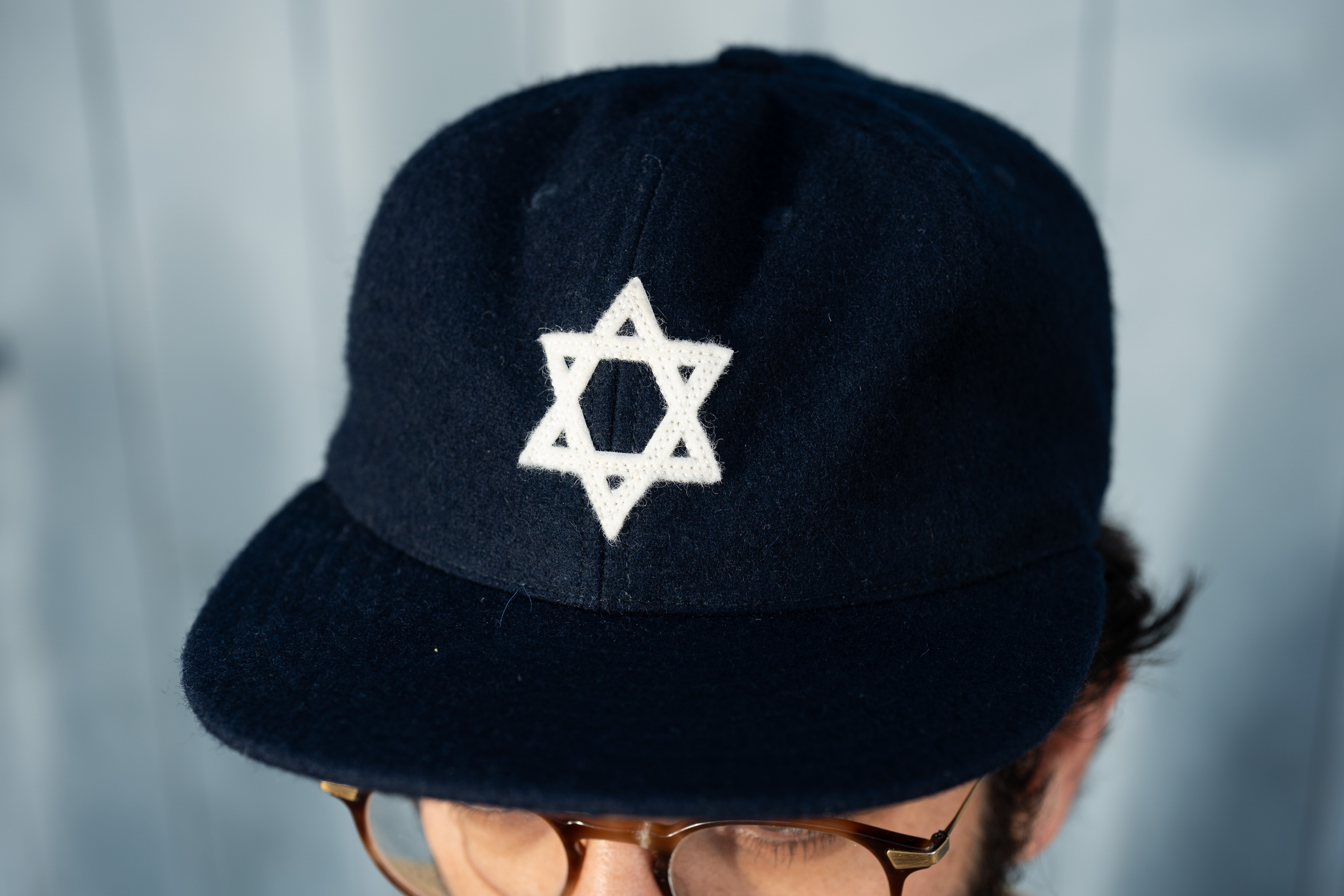 A person wears a navy blue cap with a white Star of David symbol on it. The person has glasses and is partially visible from the eyebrows up.