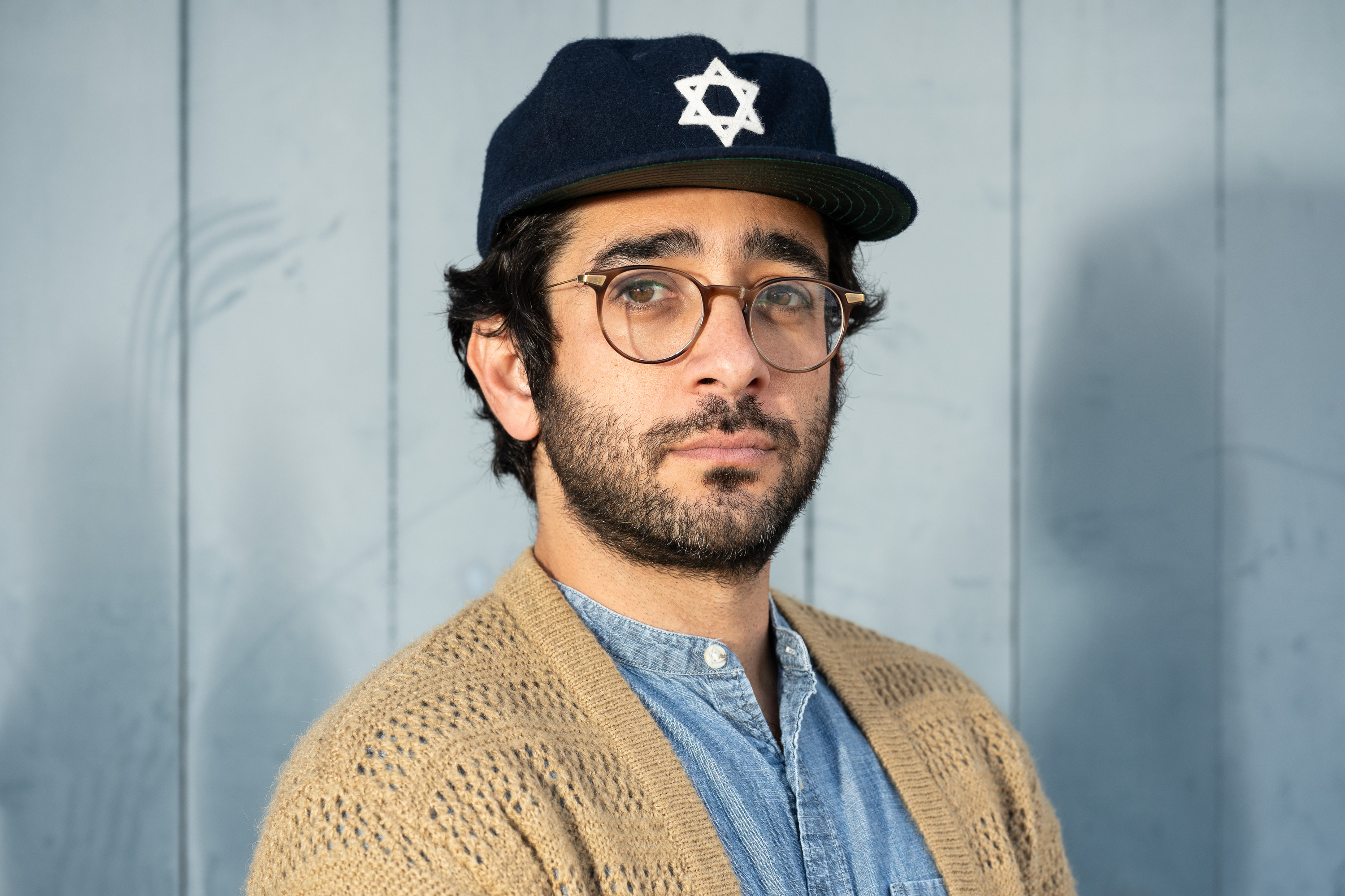 A person wearing glasses and a navy cap with a Star of David symbol is standing against a light blue wooden background, dressed in a denim shirt and a tan knit sweater.
