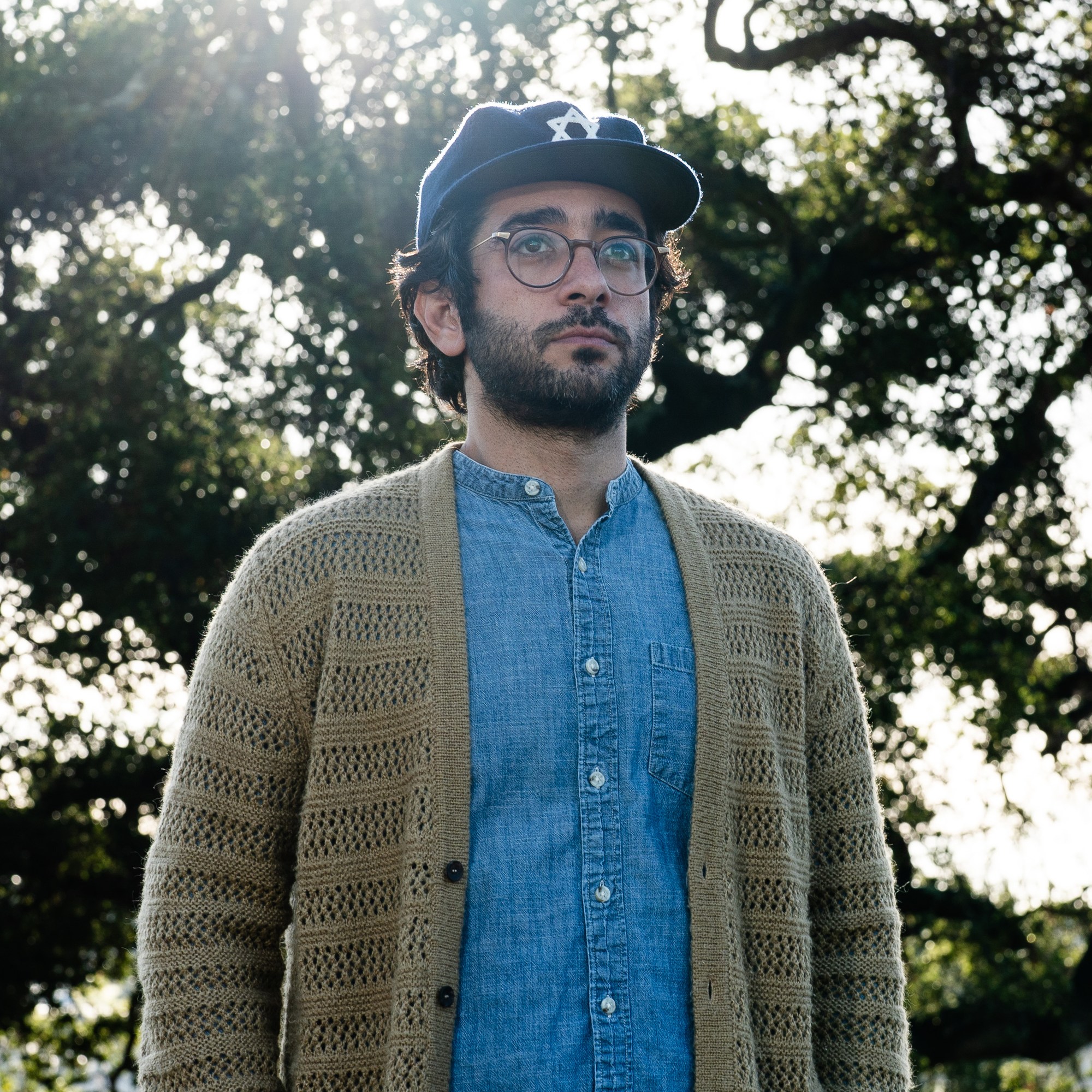 A person with a beard, glasses, and baseball cap stands outdoors. They wear a blue shirt and knitted cardigan. Sunlight filters through trees in the background.