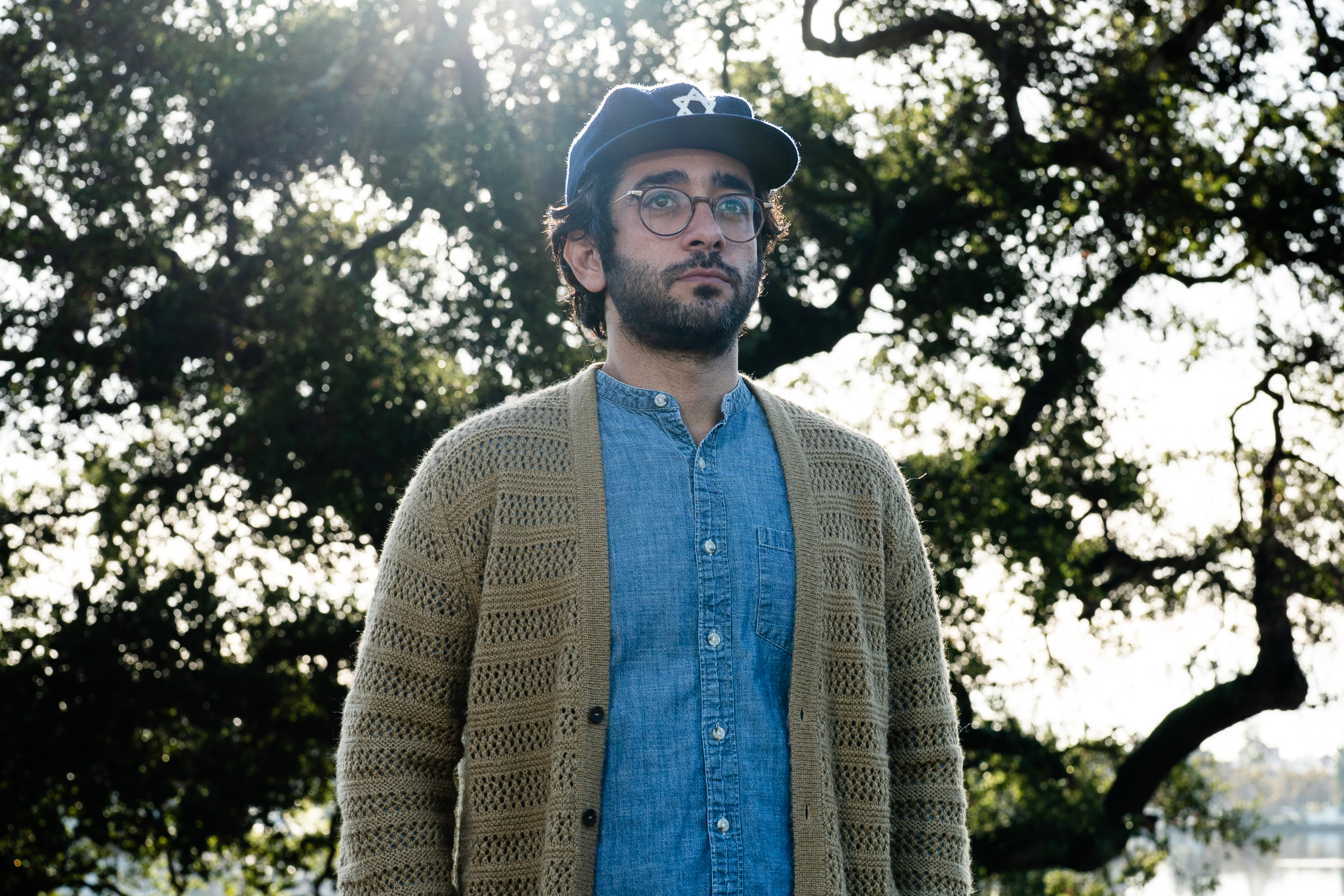 A person with a beard, glasses, and baseball cap stands outdoors. They wear a blue shirt and knitted cardigan. Sunlight filters through trees in the background.