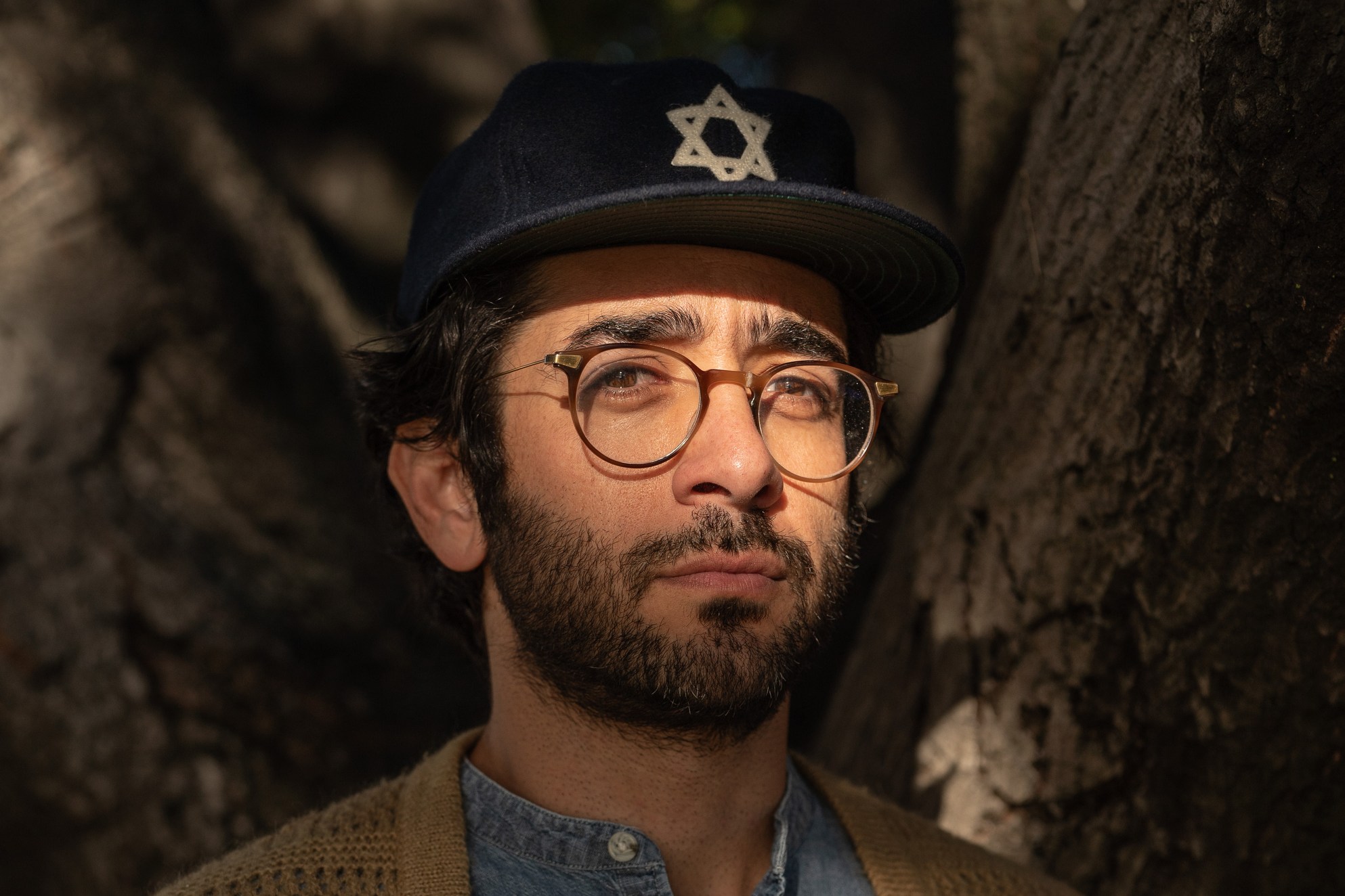 A man with glasses and a beard stands in dappled sunlight. He wears a denim shirt, beige cardigan, and a dark cap with a Star of David. Dark tree trunks frame him.