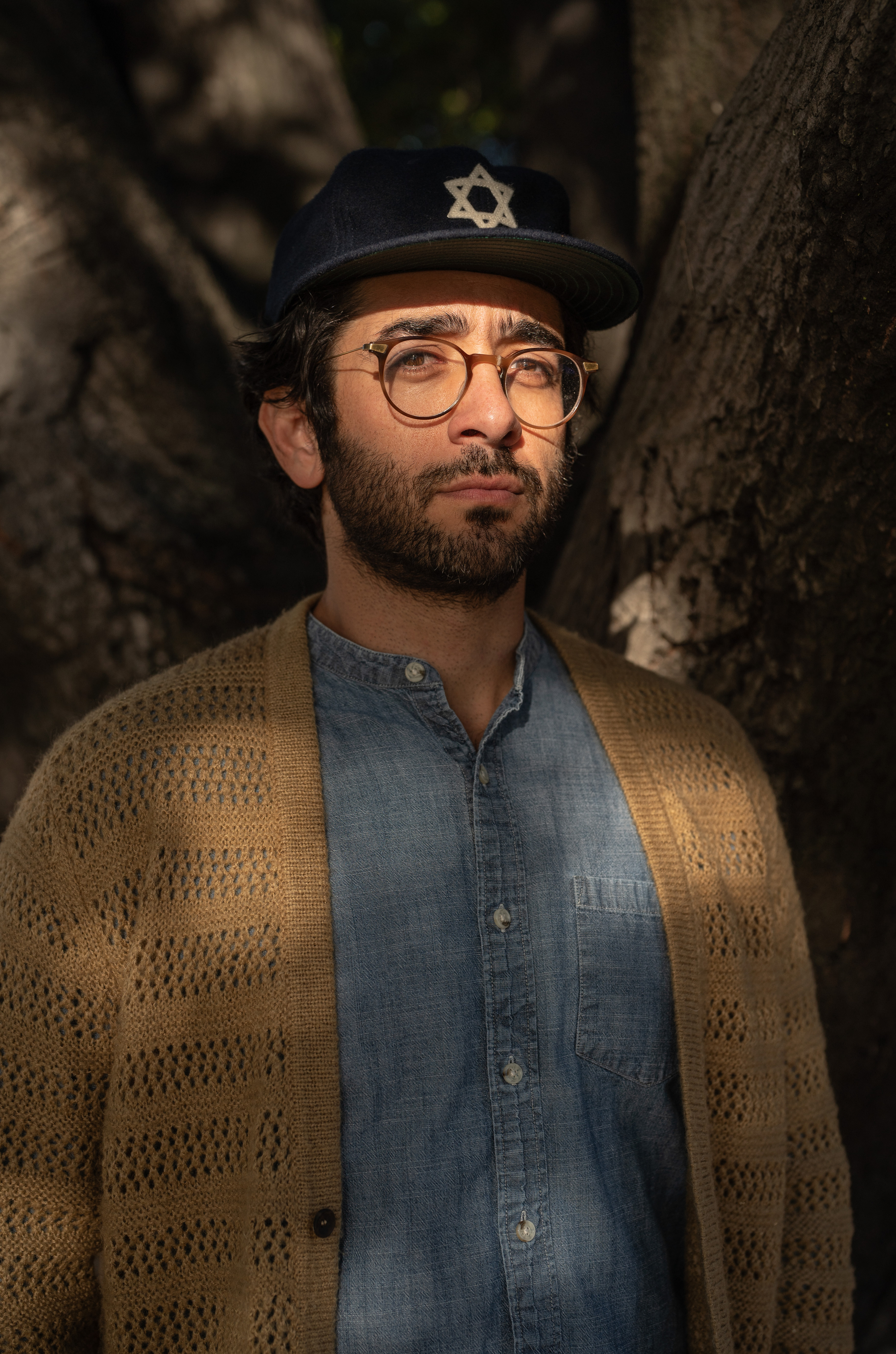 A man with glasses and a beard stands in dappled sunlight. He wears a denim shirt, beige cardigan, and a dark cap with a Star of David. Dark tree trunks frame him.