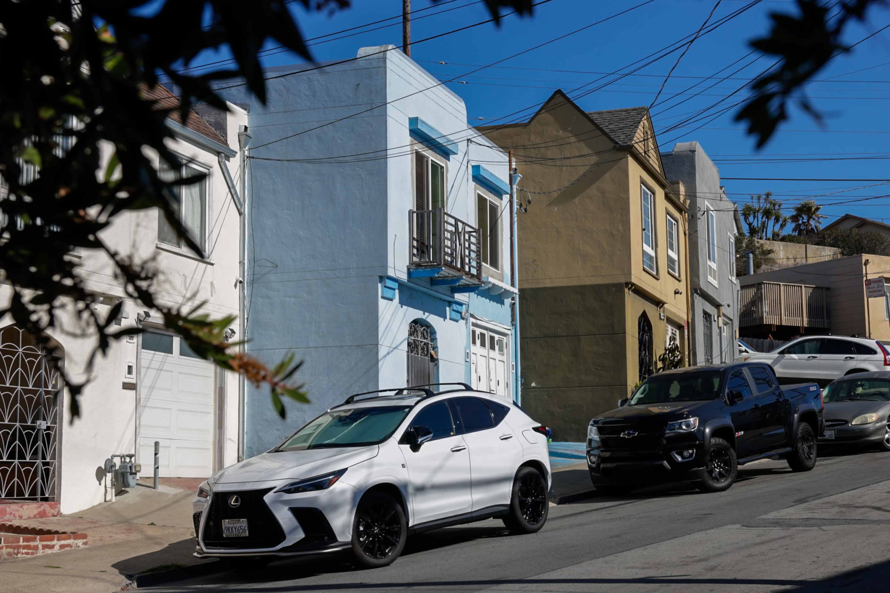 A steep street lined with colorful houses is pictured. Parked cars include a white SUV and a black pickup truck, with clear blue skies overhead.