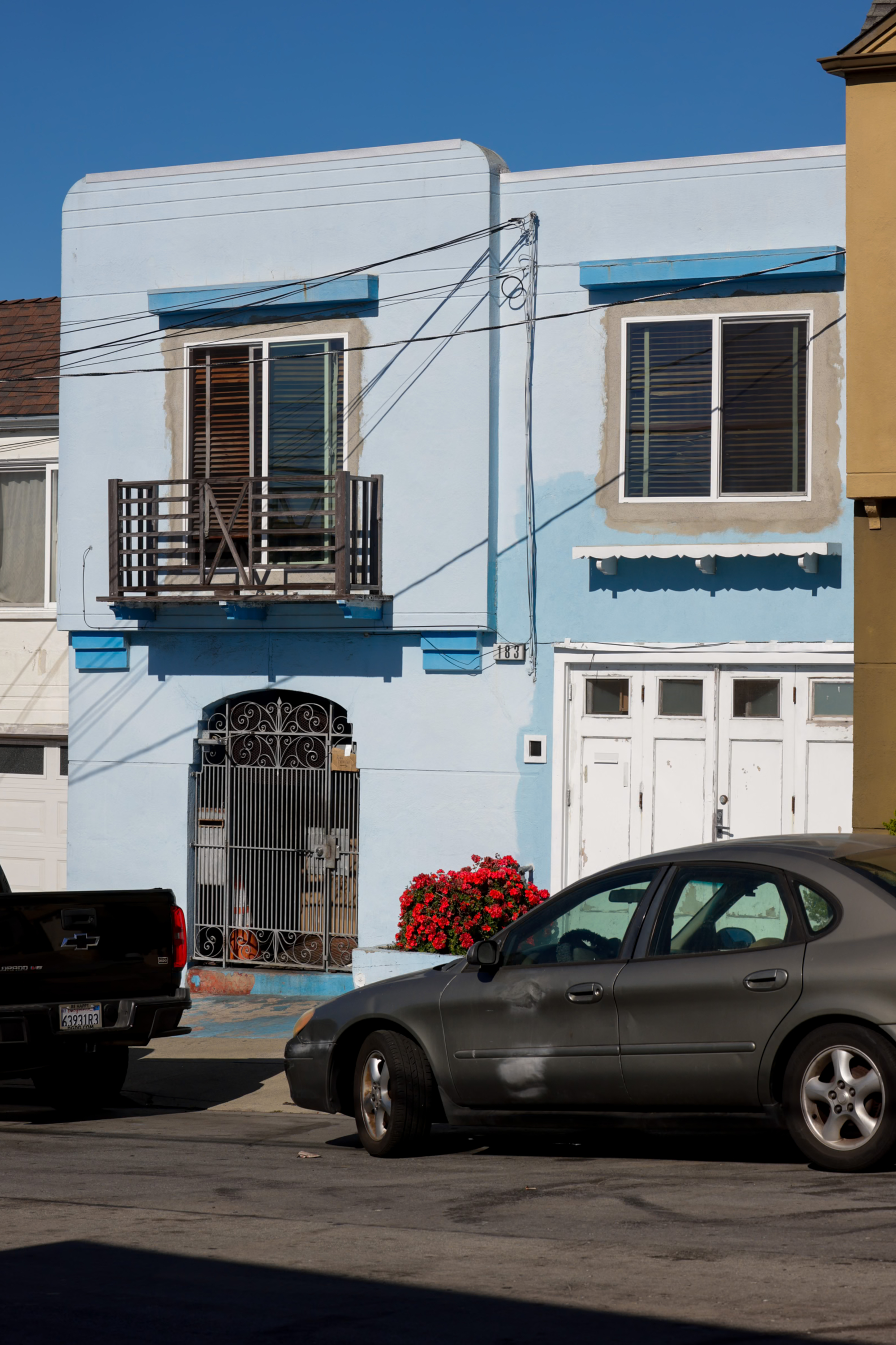 A blue two-story house with a balcony, red flowers, and two parked cars in front, one black truck, and one gray sedan with visible dents.