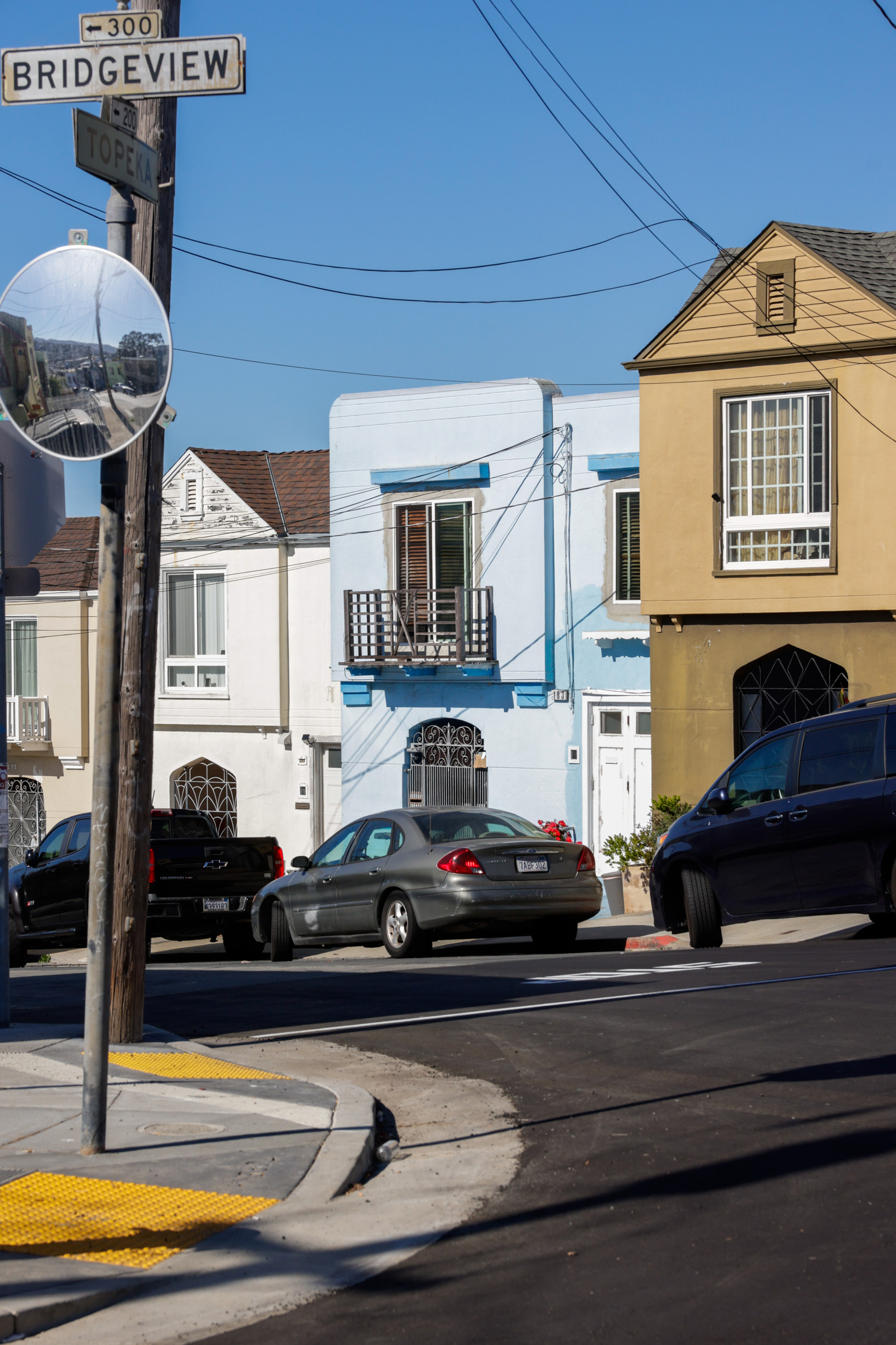 A street corner with colorful houses, parked cars, and a mirror reflecting the scene. Street signs read "Bridgeview" and "Topeka" under a clear blue sky.