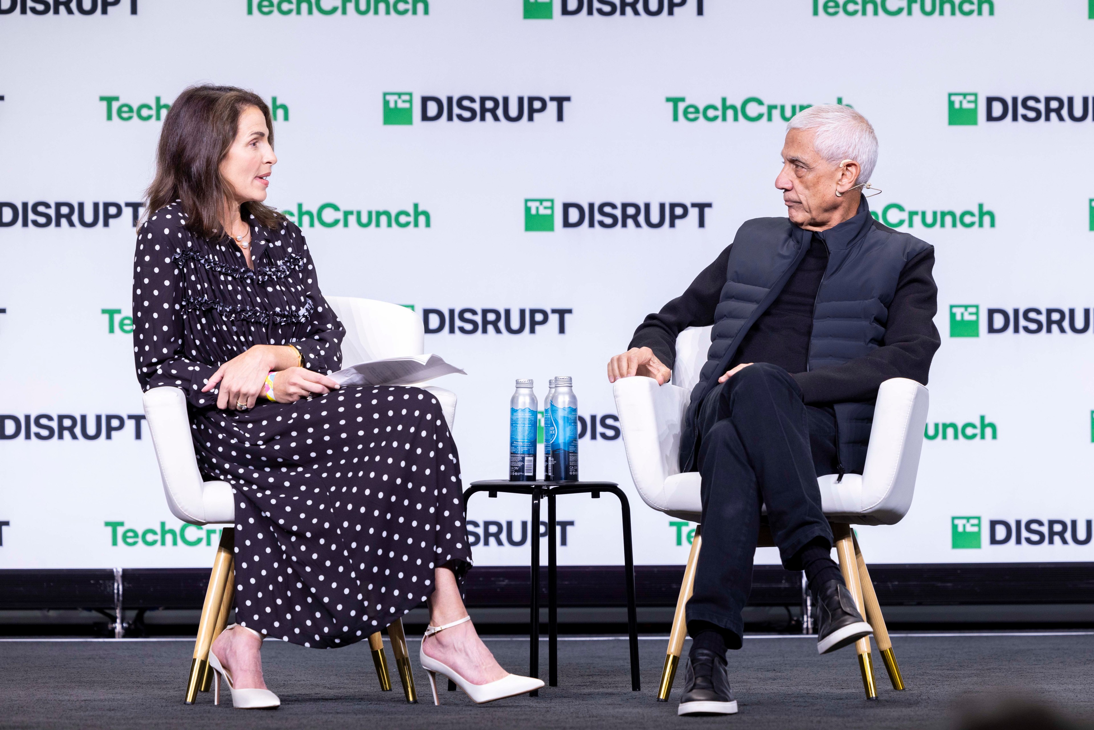 A woman in a polka dot dress and a man in a dark outfit sit on stage discussing. A table with two water bottles is between them, with &quot;TechCrunch Disrupt&quot; in the background.